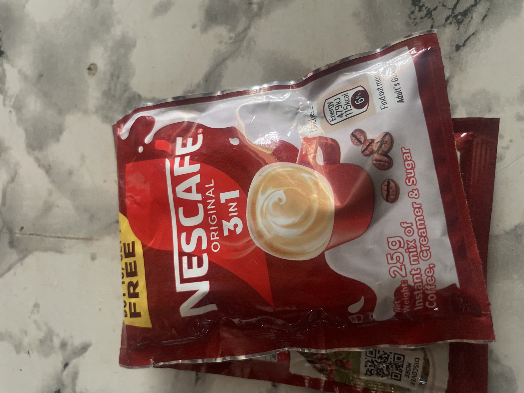 The image shows two Nescafe Original 3-in-1 instant coffee sachets on a marble surface. The top sachet is partially visible, displaying the Nescafe logo, "Original 3in1," and a depiction of a coffee cup with latte art. Nutritional information, including energy content and a percentage, is also visible. The bottom sachet is mostly obscured by the top one. A QR code and the text "DISCOVER MORE" are visible on the left side of the top sachet. The background is a light-colored marble surface. The scene is indoors and appears to be a product display or personal item. There are no people or any indication of the city of New Stadium, Nigeria.