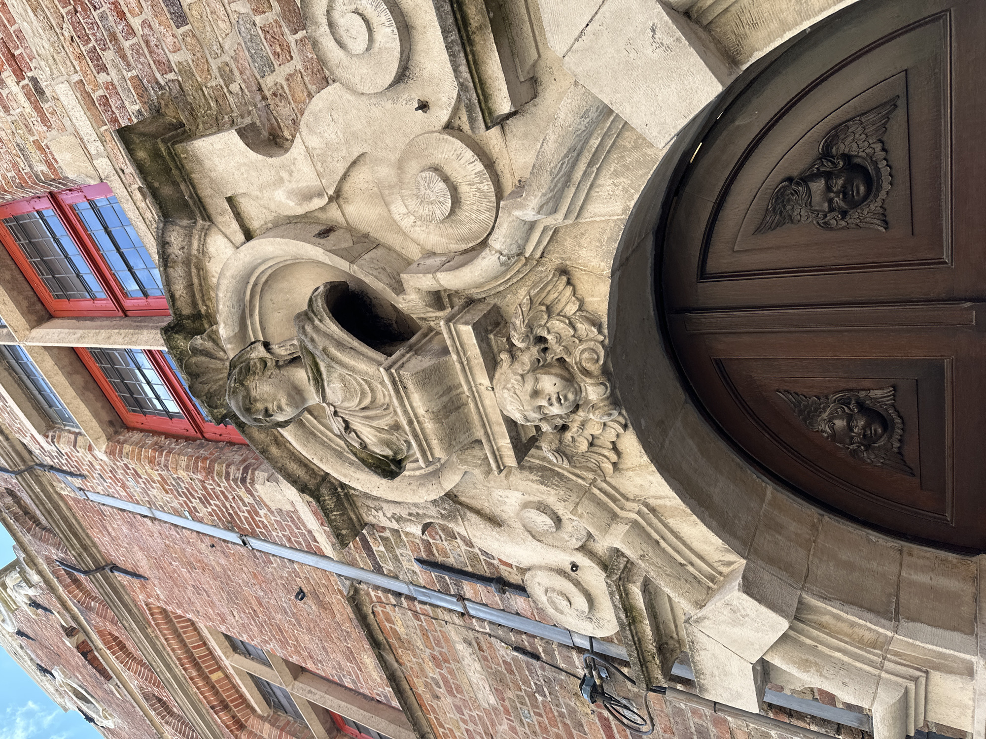 This is a low-angle, close-up, eye-level shot of the ornate facade of a historic building in Bruges, Belgium. The building's exterior is constructed primarily of red brick, with some sections of lighter-colored stone. The focal point is a detailed stone carving above a large, dark wooden door. The carving features a bust of a person at the top, with a cherub's head and wings positioned below it. The cherub's head is carved in relief and appears to be made of stone. The wooden door below the carving is divided into two panels and features two similar carved reliefs of cherubs' heads with wings. The red window frames of the building add a vibrant contrast to the brick and stone. The overall impression is one of architectural grandeur and historical detail.