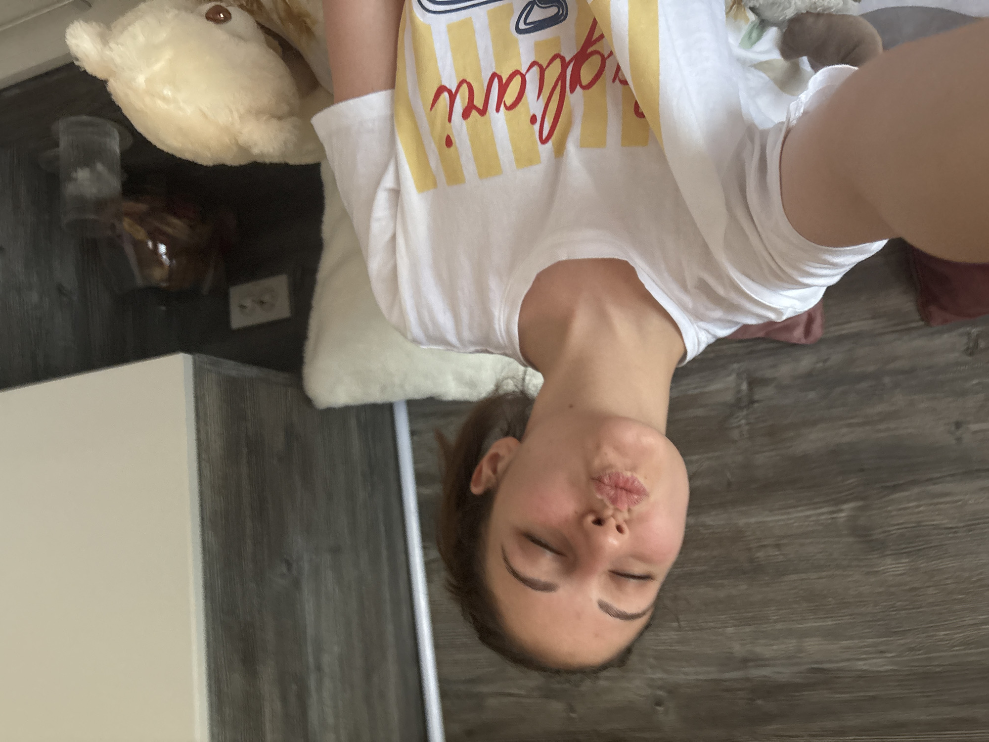 A woman with closed eyes and pursed lips is lying on a floor, taking a selfie. She has brown hair pulled back, and is wearing a white t-shirt with yellow and red stripes. To her left, there is a plush teddy bear on a white cushion and a dark table. The location is likely indoors, on a grey-toned wood floor. There is no information to support that this image was taken in Bulgaria.