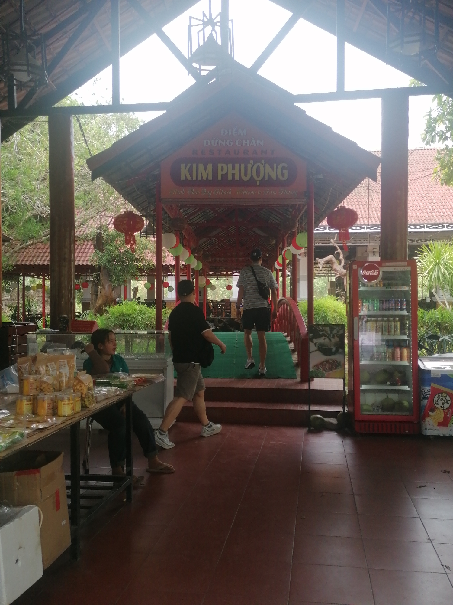 The image captures the entrance to "Kim Phuong Restaurant," a rest stop located in Nam Khanh Vinh, Vietnam. The restaurant's sign is prominently displayed. People are walking into the restaurant; one is dressed in shorts and a striped shirt, while another wears shorts and a black shirt. A seated individual is present at a table where various food products are displayed for sale. A red Coca-Cola refrigerator is placed adjacent to the entrance steps. The floor is tiled in a reddish-brown color. The restaurant is decorated with balloons and red lanterns, suggesting a celebratory or welcoming atmosphere. There is visible vegetation outside the restaurant.