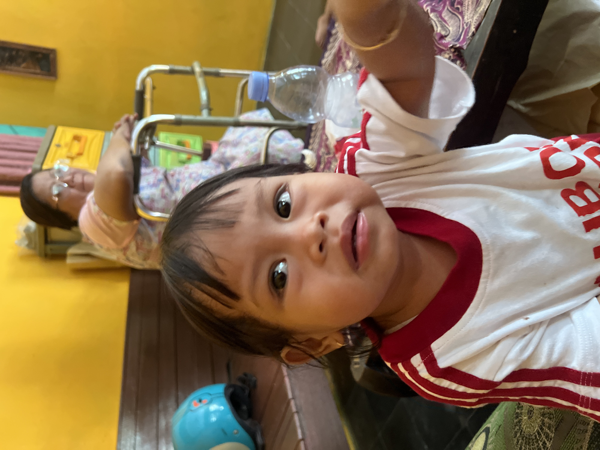 A child, likely a toddler, with dark hair and brown eyes is in the foreground, looking at the camera with a slightly distressed expression. They are wearing a white t-shirt with red trim and red stripes down the sleeves. To the left of the child, a blue helmet is visible on a wooden surface. In the background, an elderly woman wearing glasses and a patterned dress is seated in a walker. She is partially obscured by the child and the walker. A clear plastic water bottle with a blue cap is on a table behind the child. The room has yellow walls. The scene appears to be indoors, possibly in a home in Kecamatan Pati, Indonesia.