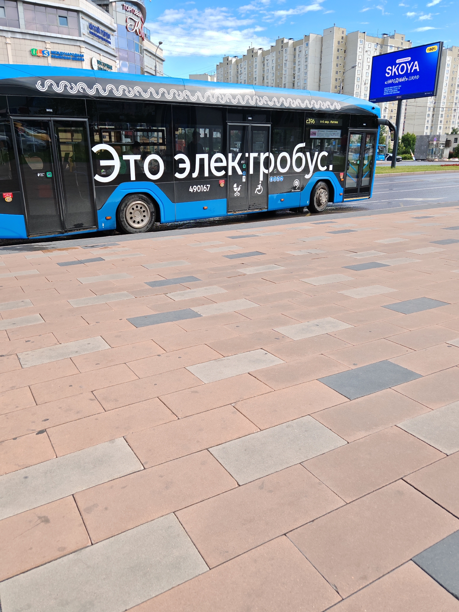 The image shows a blue and black electric bus parked on a city street. The bus has the words "Это электробус." (This is an electric bus.) written in large white letters on its side. There is also a bus number, "490167," and a route number, "c396." The bus has tinted windows and a modern design. In the background, there are several apartment buildings and a billboard advertising "SKOYA 'НАРОДНЫЙ' ШКАФ" (SKOYA 'PEOPLE'S' WARDROBE') with "80%" displayed. The street in the foreground is paved with rectangular tiles of various shades of pink and gray. The sky is blue with some white clouds, suggesting it is daytime and the weather is fair. The scene appears to be a typical urban environment. There are no people clearly visible in the image, but the bus implies public transportation is in use. The overall mood is neutral and informative, focusing on the presence of the electric bus.