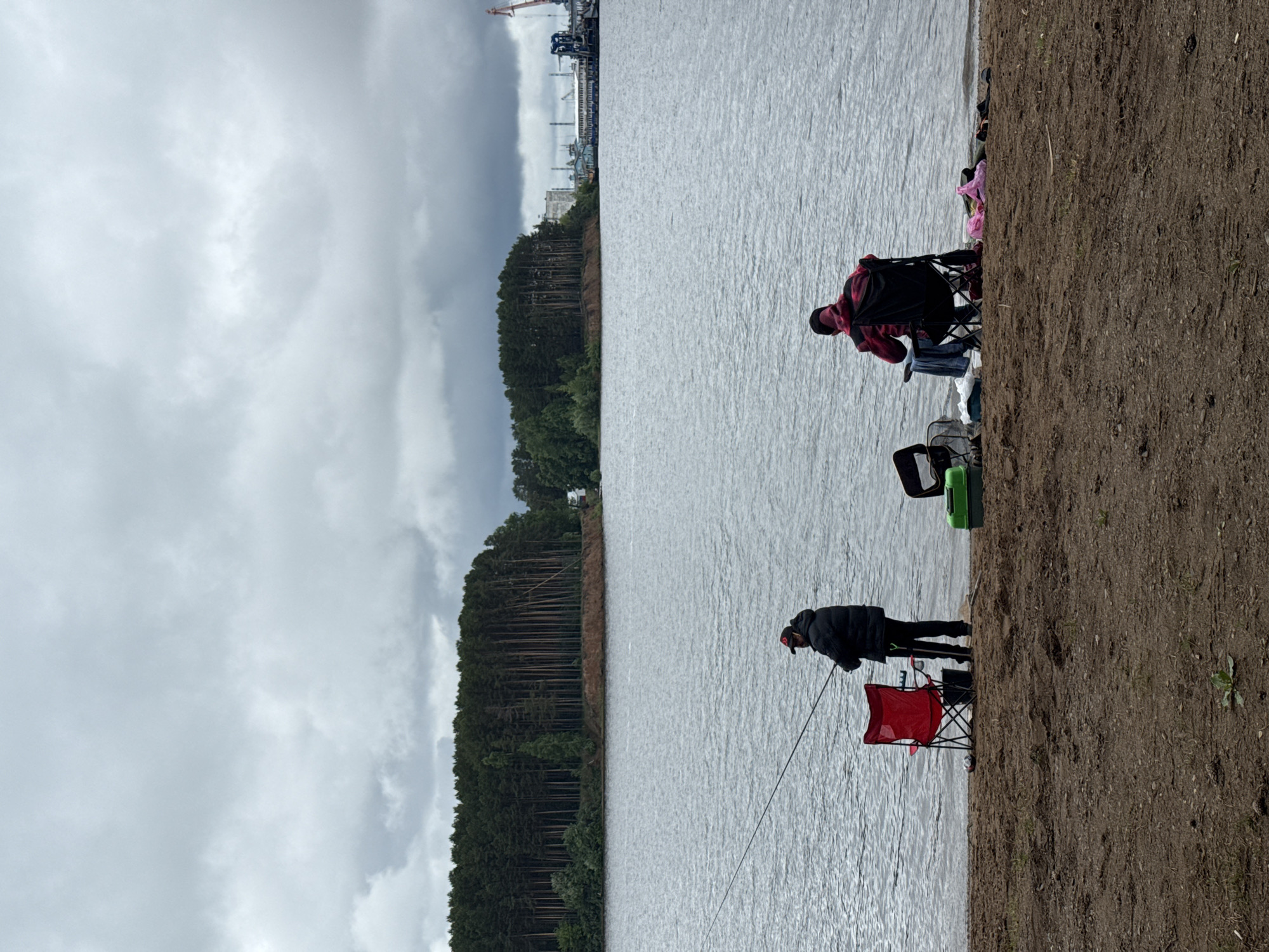 This image, located in the city of Chaikovsky, Russia, depicts a cloudy daytime scene at the edge of a large body of water. In the foreground, a sandy, reddish-brown dirt beach extends across the bottom third of the frame, showing sparse green vegetation and faint ground impressions.
Two individuals are present on the beach, engaged in fishing. The person on the left stands facing slightly right, wearing a dark puffer jacket, dark pants, and a dark baseball cap with a red emblem. This individual holds a fishing rod angled over the water. A red collapsible camping chair is positioned behind and to the left of this person, and a dark bucket is visible near their feet. The person on the right is seated in a black collapsible camping chair, facing away from the camera towards the water. This individual wears a dark jacket with a red and black plaid or camouflage pattern, dark pants, and a dark cap. Near their chair on the sand are several items, including a green tackle box, a fishing net, a blue garment, and a pink garment.
The midground is dominated by the calm, greyish-blue water, which exhibits slight ripples across its surface.
The background features the opposite bank of the water body. On the left side, a dense forest of tall, slender pine trees (conifers) rises from an elevated, visibly eroded dirt bank. To the right, beyond a break in the trees, an industrial area is present, characterized by several buildings, multiple cranes (including a prominent red and white gantry crane), and large vessels or barges docked along the shore.
The sky occupies the upper portion of the image and is completely overcast with thick, uniform grey clouds, indicating a lack of direct sunlight.