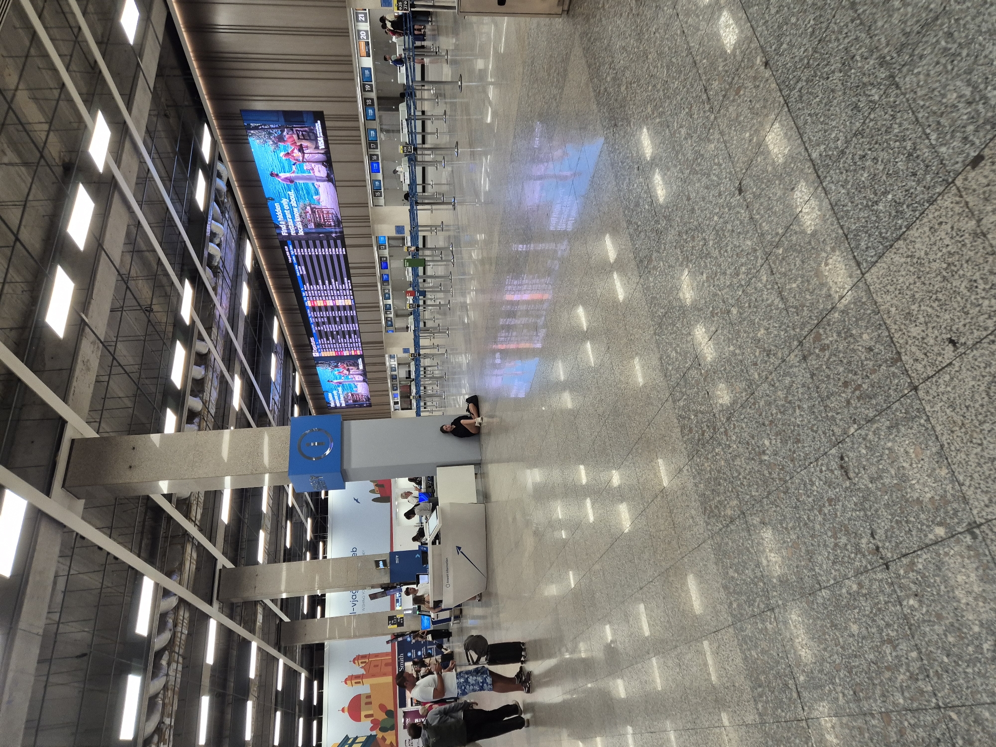 The image depicts the interior of an airport terminal, likely the passenger departure area, in Luqa, Malta. The scene is characterized by a vast, polished granite floor reflecting the overhead lighting and large digital display screens. Several people are visible, some standing and interacting at a customer service desk, while others are seated or walking with luggage. A woman is seen sitting on the floor, leaning against a pillar, near the information desk. The departure board displays flight information, and queues of people are absent from the check-in counters, suggesting a less busy period or an area not yet in full operation. The architectural style includes large concrete pillars supporting a high ceiling with numerous fluorescent lights. The overall impression is one of modern airport infrastructure.