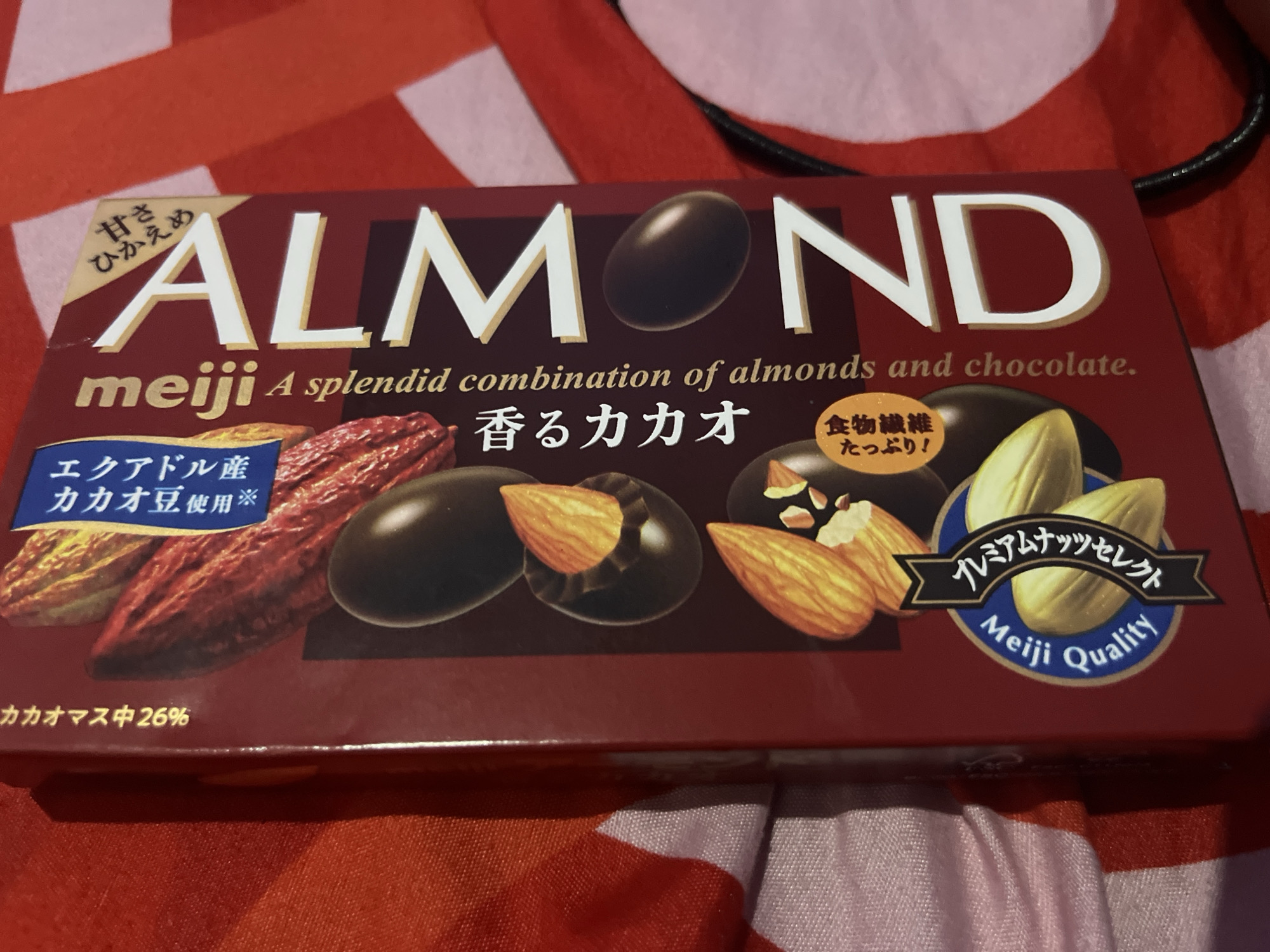 The image shows a close-up of a Meiji Almond chocolate box. The box is red with the brand name "ALMOND" prominently displayed in large, white, stylized letters. Below the main title, it reads "meiji A splendid combination of almonds and chocolate." There are visual depictions of chocolate-covered almonds and whole almonds on the box. Text in Japanese characters is also present, including "甘さひかえめ" (slightly sweet) and "香るカカオ" (fragrant cacao). A smaller circular badge reads "プレミアムナッツセレクト Meiji Quality" (Premium Nut Select Meiji Quality). The box also indicates "カカオマス中26%" (26% cacao mass). The background appears to be a red and white striped fabric. There is no indication or visual information related to the city of Boja, Indonesia, in this image.