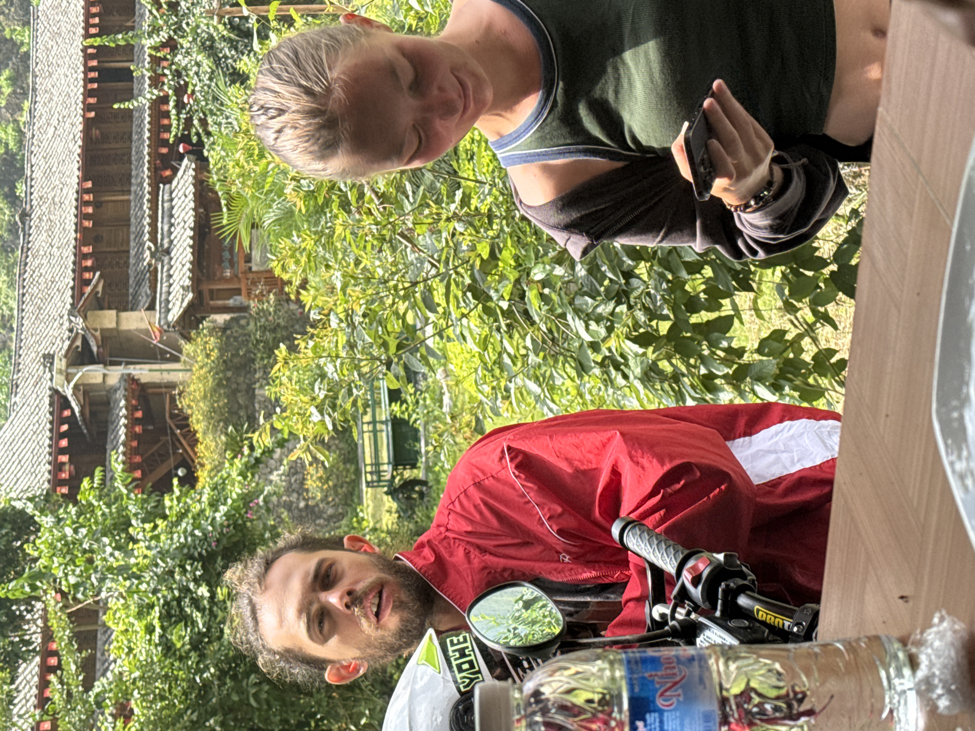 Two people, a man and a woman, are pictured outdoors with a motorcycle. The man is sitting on the motorcycle, wearing a red jacket, with a helmet in front of him. A water bottle sits on the motorcycle. The woman is standing next to him, looking at a smartphone she holds in her hand. Behind them are trees and foliage, and a traditional-style building with tiered roofs. The image suggests the scene is in a location like the Mèo Vạc Commune in Vietnam, characterized by traditional architecture and natural surroundings.