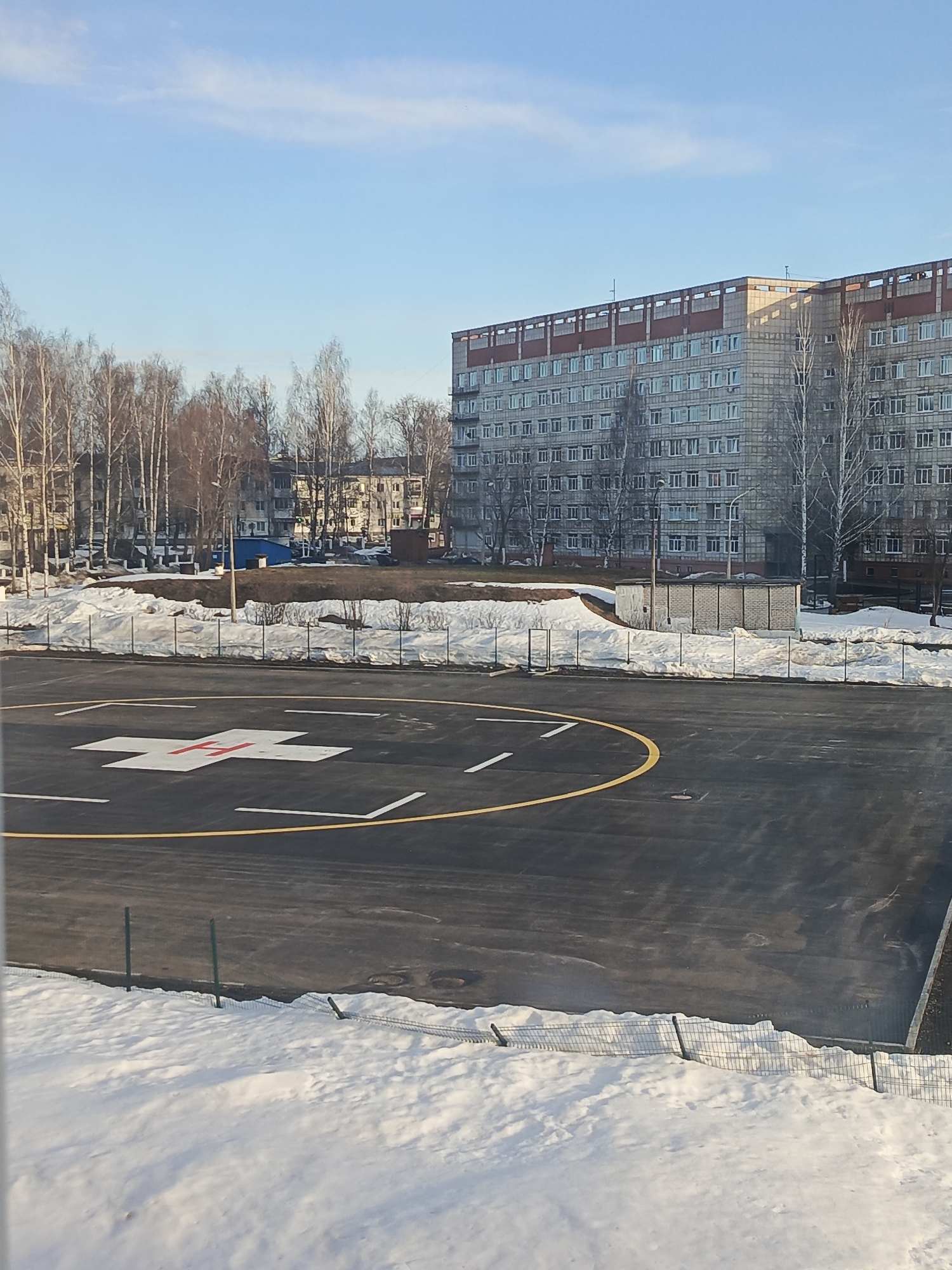 The image displays an outdoor scene in Chaikovsky, Russia, likely during winter or early spring, under a clear blue sky. The foreground features a helipad, a dark asphalt surface marked with a central white 'H' overlaid by a red cross, encircled by a yellow line and segmented white dashes. A low, wire mesh fence separates the helipad from a snow-covered area in the immediate foreground. Beyond the helipad, accumulated snow covers the ground, with scattered bare deciduous trees visible. In the midground, a small, light-colored brick utility building stands amidst the snow and trees. The background is dominated by a large, multi-story building, constructed from light-colored masonry blocks with a reddish-brown upper perimeter. This building features numerous evenly spaced windows across its facade. No people, vehicles, or active events are present within the frame.