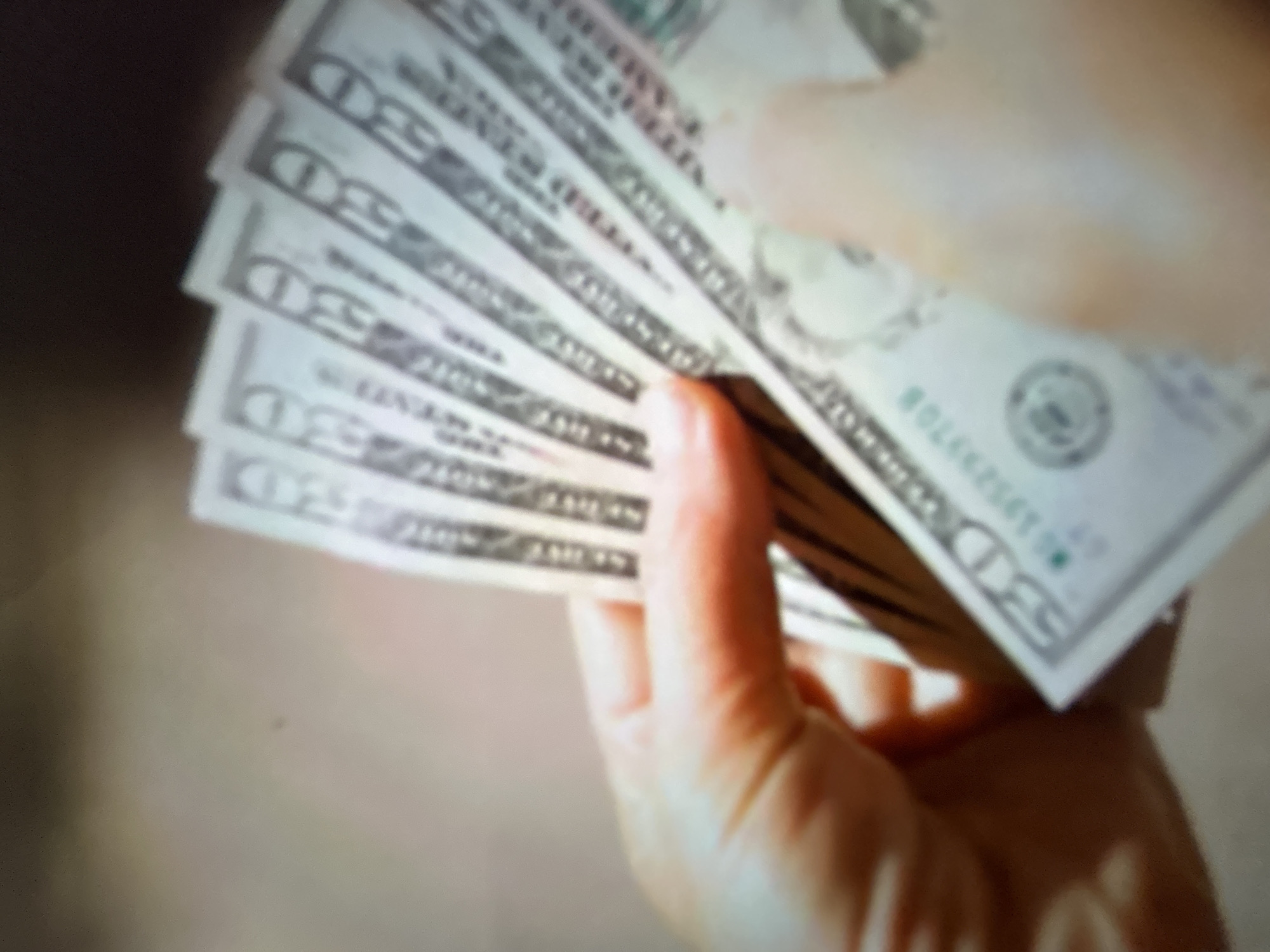This media file shows a close-up, blurred view of a person's hands holding a fan of US dollar bills. The hands are light-skinned and appear to be cupping the money. The bills are mostly visible on the left side of the frame, fanned out and held by fingers. The edges of several bills are visible, showing the characteristic green and white design of US currency. Some text is discernible on the bills, including the number "50" appearing multiple times, suggesting they are fifty-dollar bills. A serial number, "B01252970B", is also partially visible on one of the bills. The background is a blurred, indistinct dark area on the left and a lighter, also blurred area on the right, giving no specific environmental clues. The lighting is soft, and there are no indications of time of day or weather. The overall impression is one of handling money, though the specific context or emotion is not clearly conveyed due to the blurriness. The location context of Gelendzhik, Russia, is provided, but not visually evident in the image.