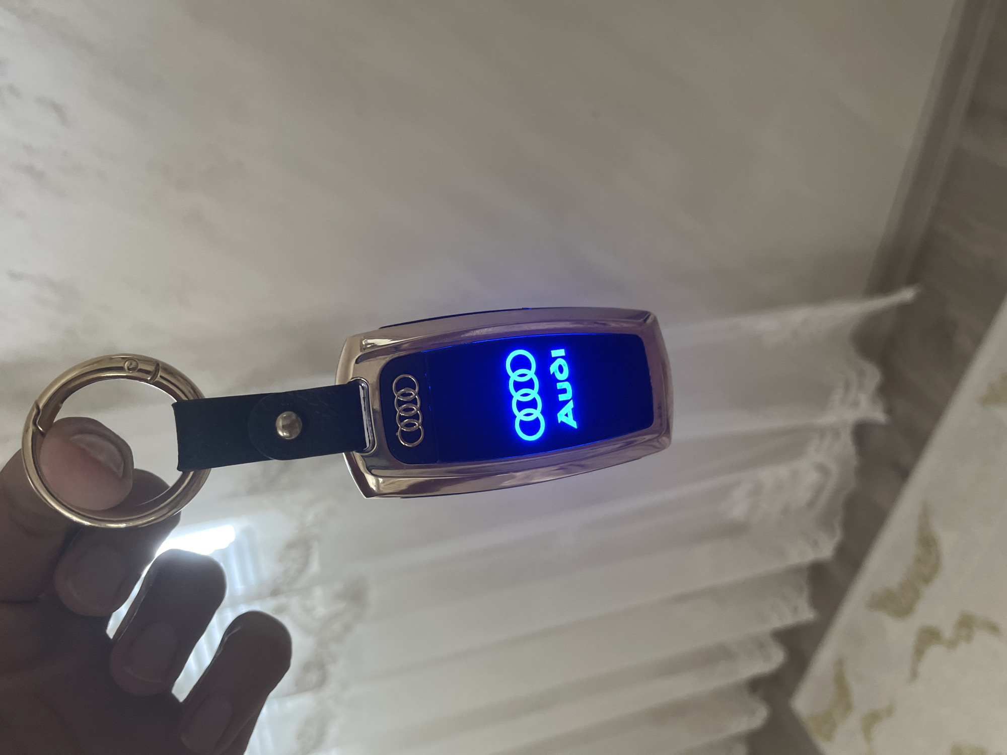 A person's hand holds up an illuminated Audi keychain in a close-up, slightly tilted shot. The keychain features a metallic, gold-toned casing with rounded edges. A black strap connects the casing to a gold-toned, circular clasp which is held by the individual's fingers and thumb. The central display panel of the keychain glows brightly with a vibrant blue light, showcasing the iconic Audi logo of four interlocking rings, accompanied by the word "Audi" printed vertically in a stylized font.
The background is an indoor setting, primarily composed of light-colored, vertically pleated curtains or blinds, suggesting a window. A plain, light-toned wall or ceiling is visible above the curtains, and to the right, a hint of a light brown surface, possibly a wooden floor or furniture, can be seen. The overall lighting appears to be subdued, allowing the blue illumination of the keychain to be prominent. The image focuses on presenting the branded accessory. This scene is located indoors in Ihtiman, Bulgaria.