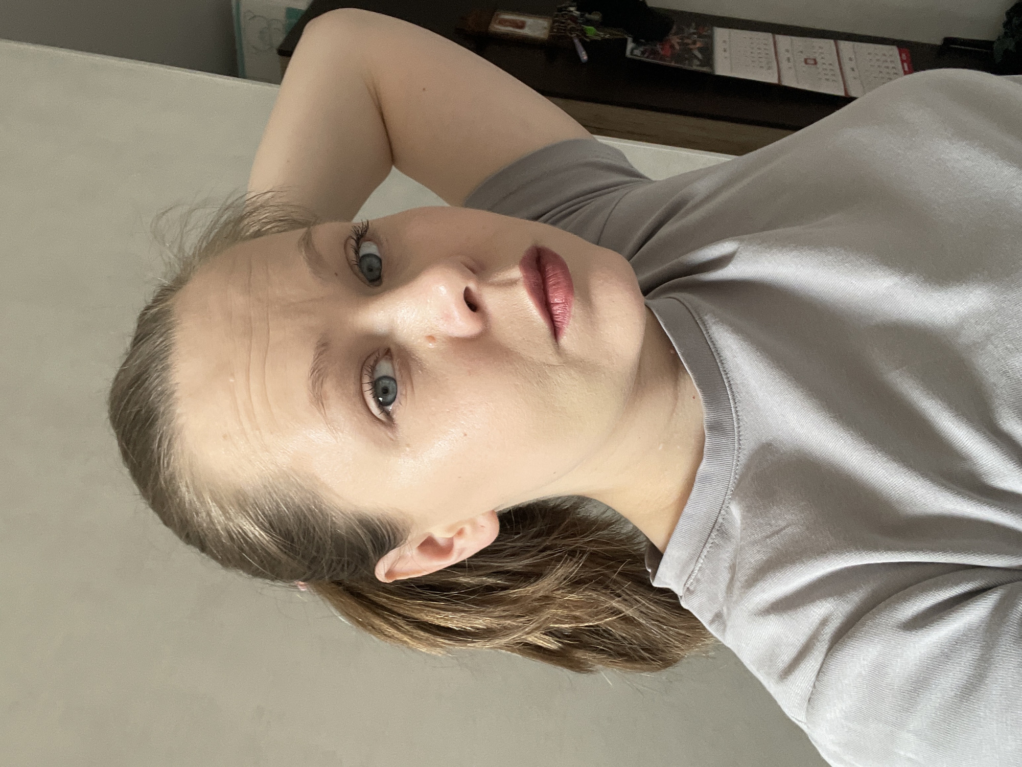 This self-portrait captures an individual, likely female, with fair skin, blue-grey eyes, and light brown hair tied back, gazing directly at the camera with a neutral expression. She is wearing a light grey short-sleeved t-shirt. The image is taken from an angle, suggesting she is lying down or reclining, with her left arm visible and bent, seemingly supporting her head.
The background points to an indoor, domestic setting in Chaikovsky, Russia. A plain, light-colored wall or surface is visible on one side, while a darker wooden shelf or piece of furniture occupies the other side. On this shelf, a calendar with grid patterns, along with a set of keys and other small, indistinct items, can be seen. The lighting is bright and even, consistent with daytime indoor conditions. No specific text is clearly legible other than the general format of a calendar.
