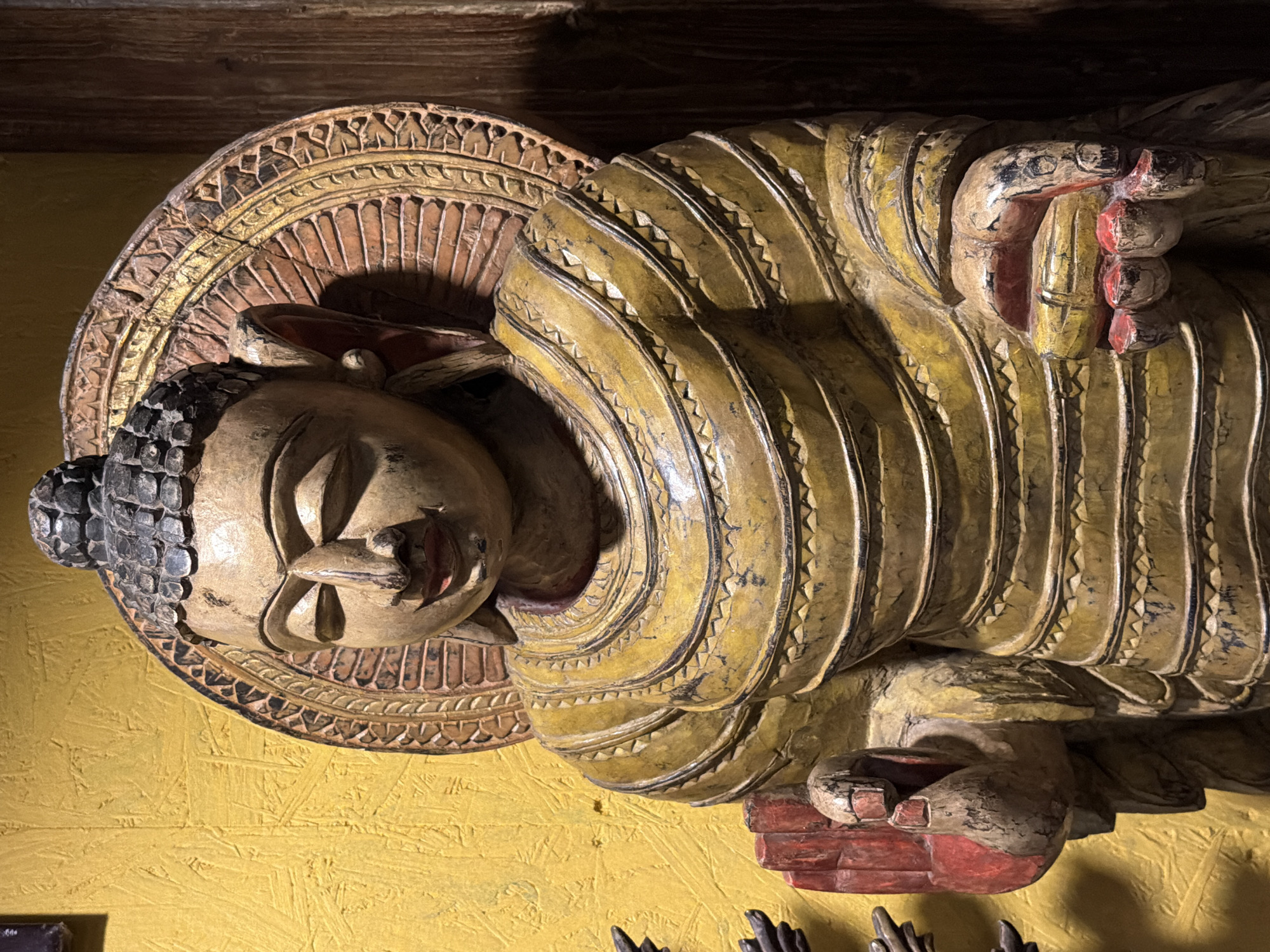 A close-up, rotated image depicts a wooden sculpture of a reclining Buddha, likely of Southeast Asian origin. The sculpture is painted, with a yellow robe featuring a repeating triangular pattern along the edges. The Buddha has black hair depicted as small, rounded shapes. The sculpture is mounted on a yellow wall, possibly a decorative wood panel. The presence of the sculpture in this setting doesn't confirm it is in Moscow, Russia, without additional contextual cues. Additional sculptures appear at the bottom. The lighting suggests an indoor environment.