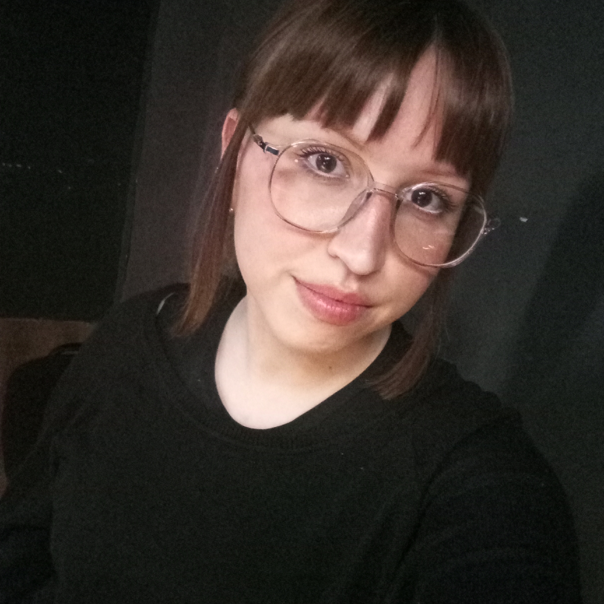 A medium indoor shot shows a fair-skinned young woman with brown hair and bangs, wearing eyeglasses with round, metal frames, and a black sweater. She is looking directly at the camera with a slight smile. The background is dark and indistinct. The image does not show any elements that can be geolocated to Chaikovsky, Russia. There is no basis to infer a location.
