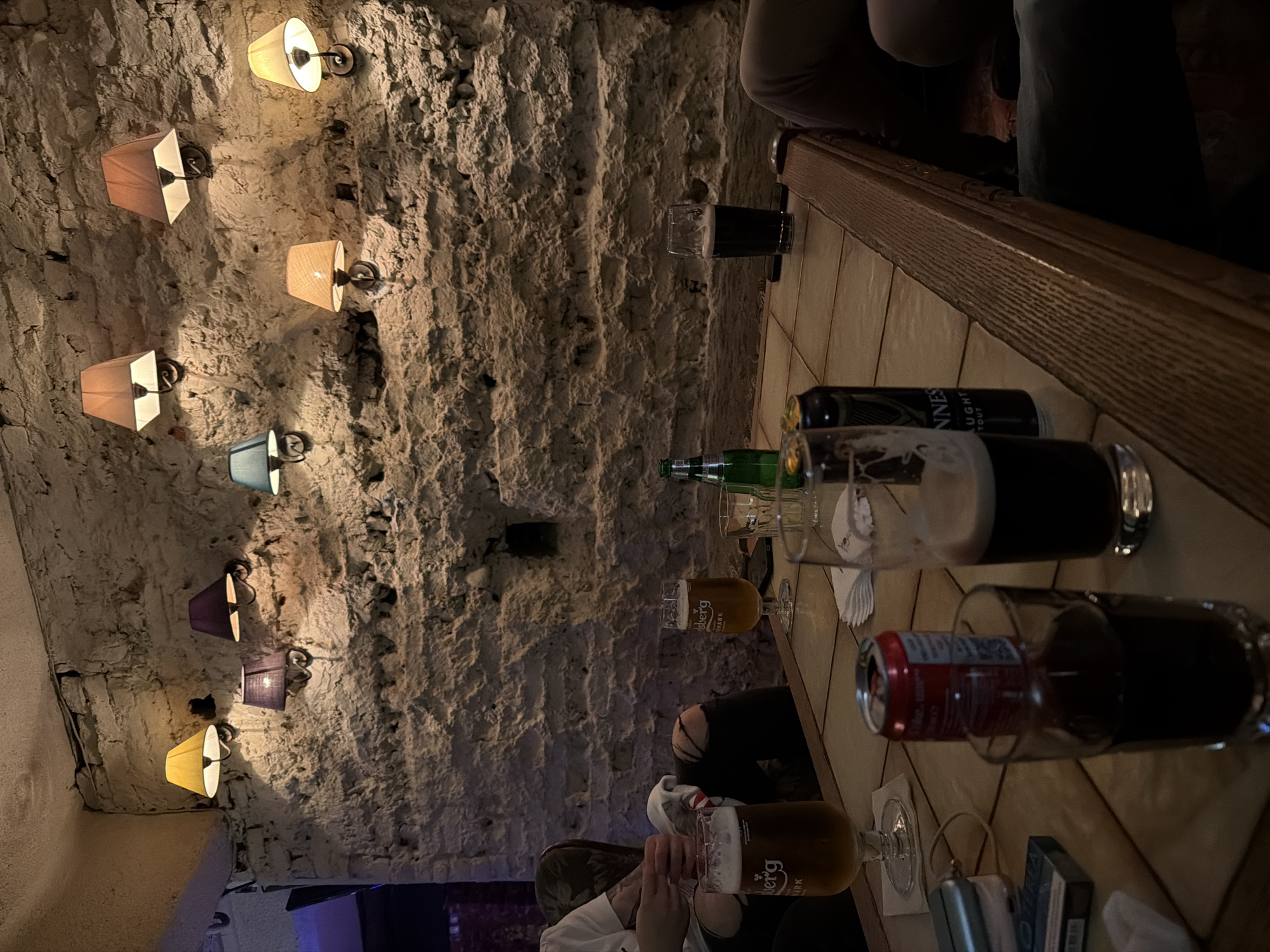 The scene depicts the interior of a restaurant or bar, characterized by a rustic aesthetic. The walls are constructed from roughly hewn stone and are adorned with a series of lamps fitted with different colored lampshades. A wooden table extends across the scene, populated with an assortment of beverages including beer glasses, bottles, and cans. The lighting within the establishment is dim, creating a warm and intimate ambiance. Patrons are seated at the table, partially visible in the frame, and appear to be engaged in casual conversation. No further details about the location in Sofia, Bulgaria, can be discerned.