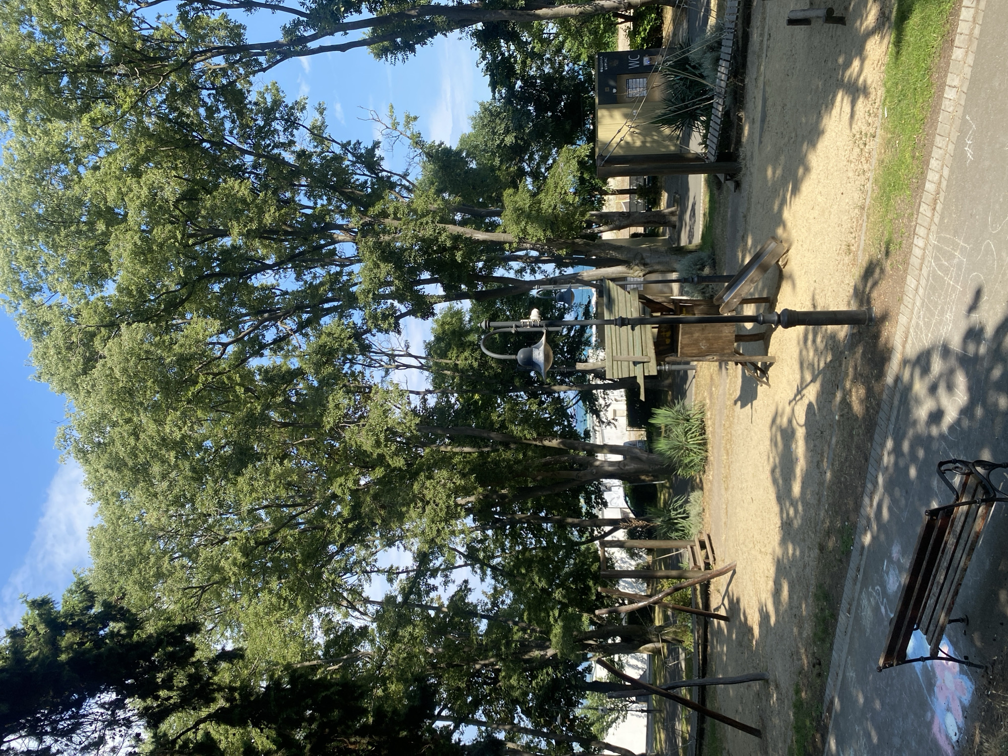 The image shows a playground area in Burgas, Bulgaria, set amongst tall, leafy trees. In the foreground, a weathered wooden bench sits on a paved pathway adorned with chalk drawings. The playground itself features a sandy surface with various wooden play structures, including a swing set and a small slide. In the background, there's a glimpse of the sea and some buildings. The overall atmosphere is one of a sunny, serene public space.
