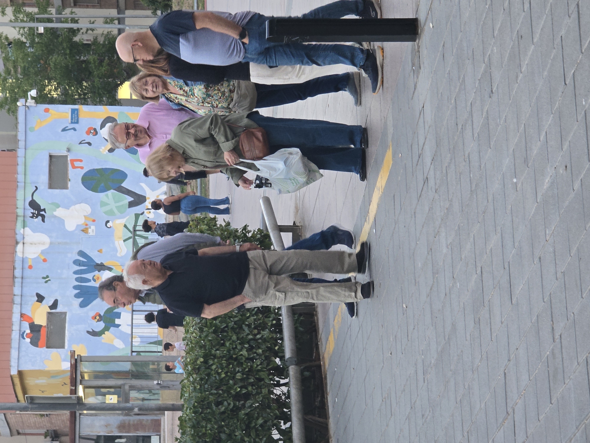 A group of at least 10 individuals stands on a gray brick sidewalk, adjacent to a yellow painted line. In the background, a building is adorned with a whimsical mural featuring cartoonish figures and shapes in shades of blue, green, and white. Several people appear to be looking at the mural, while others stand near a metal railing. The scene is located in Cornellà de Llobregat, Spain.