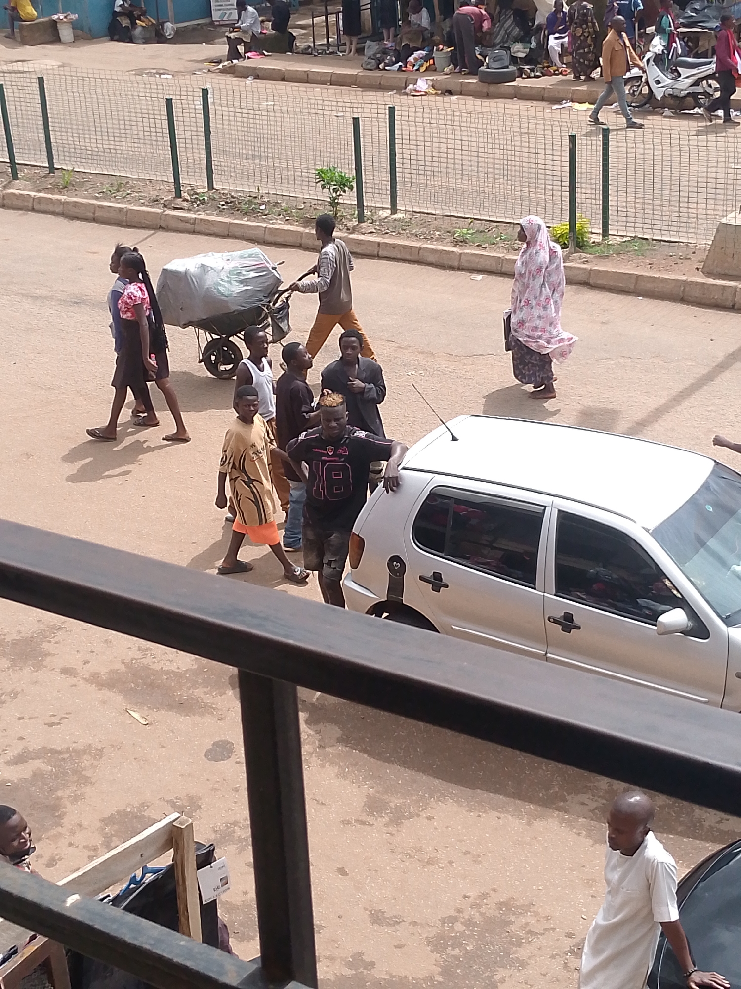 This aerial, natural-light photograph captures a street scene in Bukuru, Nigeria. A group of people are visible on a dusty, paved road. In the foreground, a young man with blonde-tipped hair, wearing a black t-shirt with the number "18" and black shorts, stands with his hands on his hips, facing the camera. To his left, a boy in a tan shirt and orange shorts walks towards the camera. Behind them, a man pushes a wheelbarrow loaded with a large, grey, plastic-wrapped item. Several other people, including women and children, are walking or standing in the mid-ground. One woman, in the upper right quadrant, is wearing a pink and white floral pattern headscarf and a dark skirt. A silver hatchback car is parked on the right side of the road, its passenger side visible. In the background, a white motorcycle is parked near a curb, and a fence made of wire mesh runs along the side of the road. The environment appears to be an urban or semi-urban area with some vegetation along the roadside. The overall impression is a busy street with pedestrian and vehicle activity.