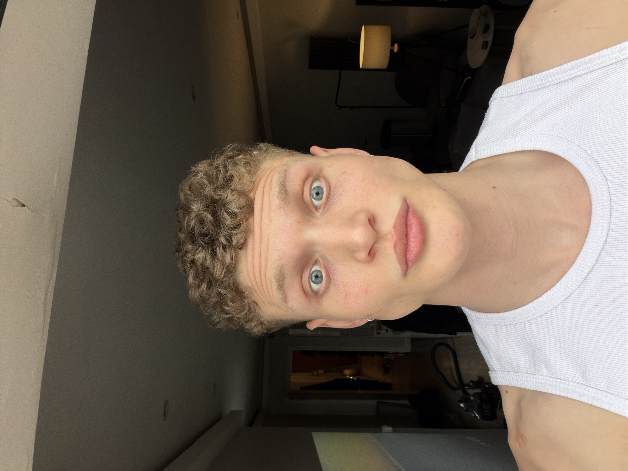 A fair-skinned male with curly blonde hair and blue eyes is positioned in the foreground, wearing a white tank top. The setting appears to be indoors, with a dimly lit room featuring a floor lamp in the background. A portion of a wall is visible on the left. The subject is looking directly at the camera. The location is not apparent.