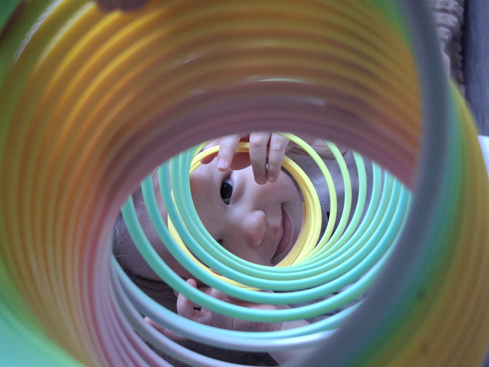 The image depicts a close-up view through a multi-colored plastic slinky toy. A child's face is visible within the center of the slinky's coils. The child's eye is directed towards the camera. The child's hands are partially visible, holding the slinky. The background is blurred. The location and city are unknown.