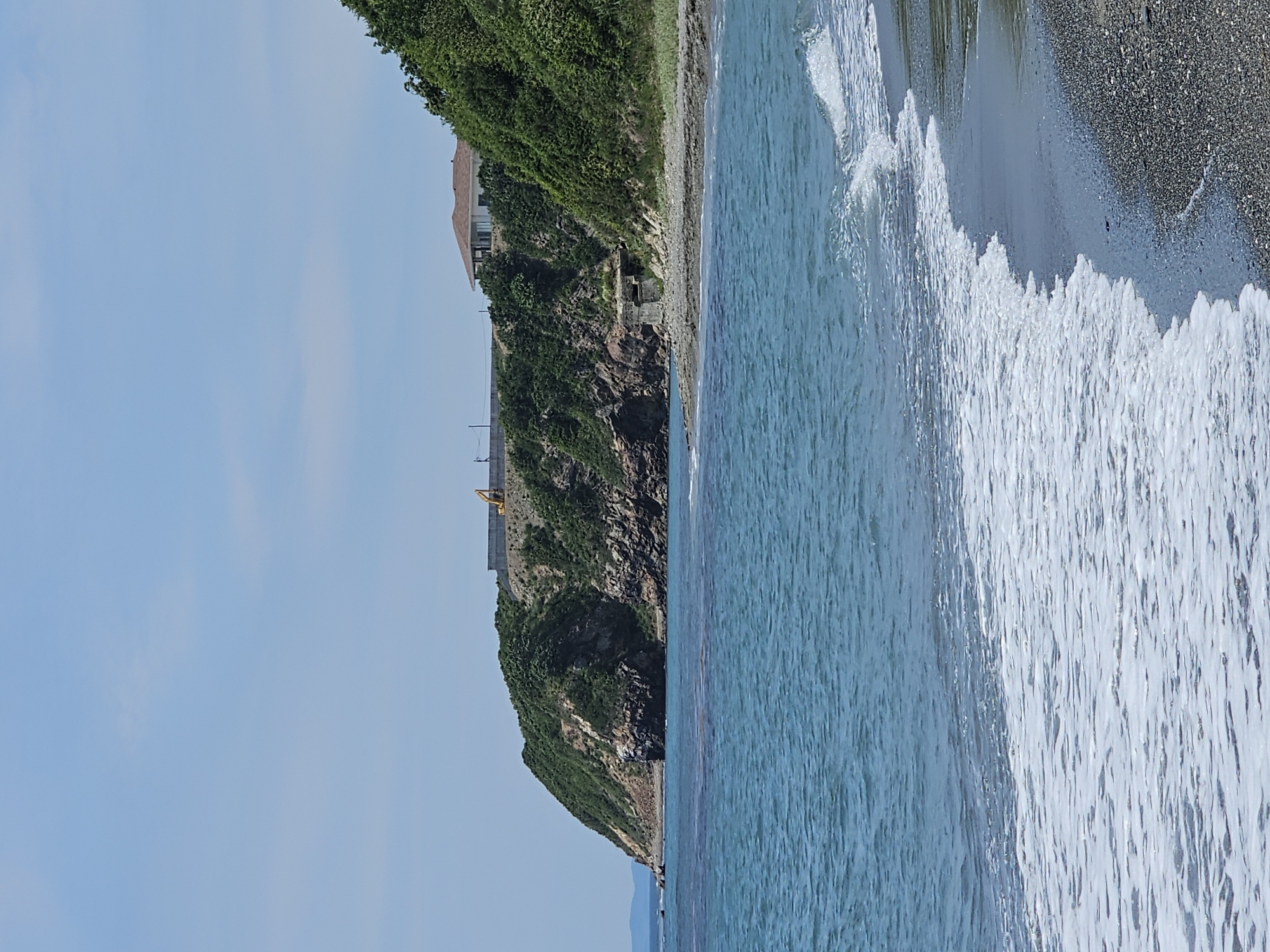 A scenic view of a rocky coastline with turquoise waters lapping onto a pebble beach. On top of the cliff, a white house with a brown roof is visible, along with a yellow construction vehicle and a concrete barrier. The sky is clear and blue with a few wispy clouds. This scene is located at the Cape of Lisy, Nakhodka, Russia.