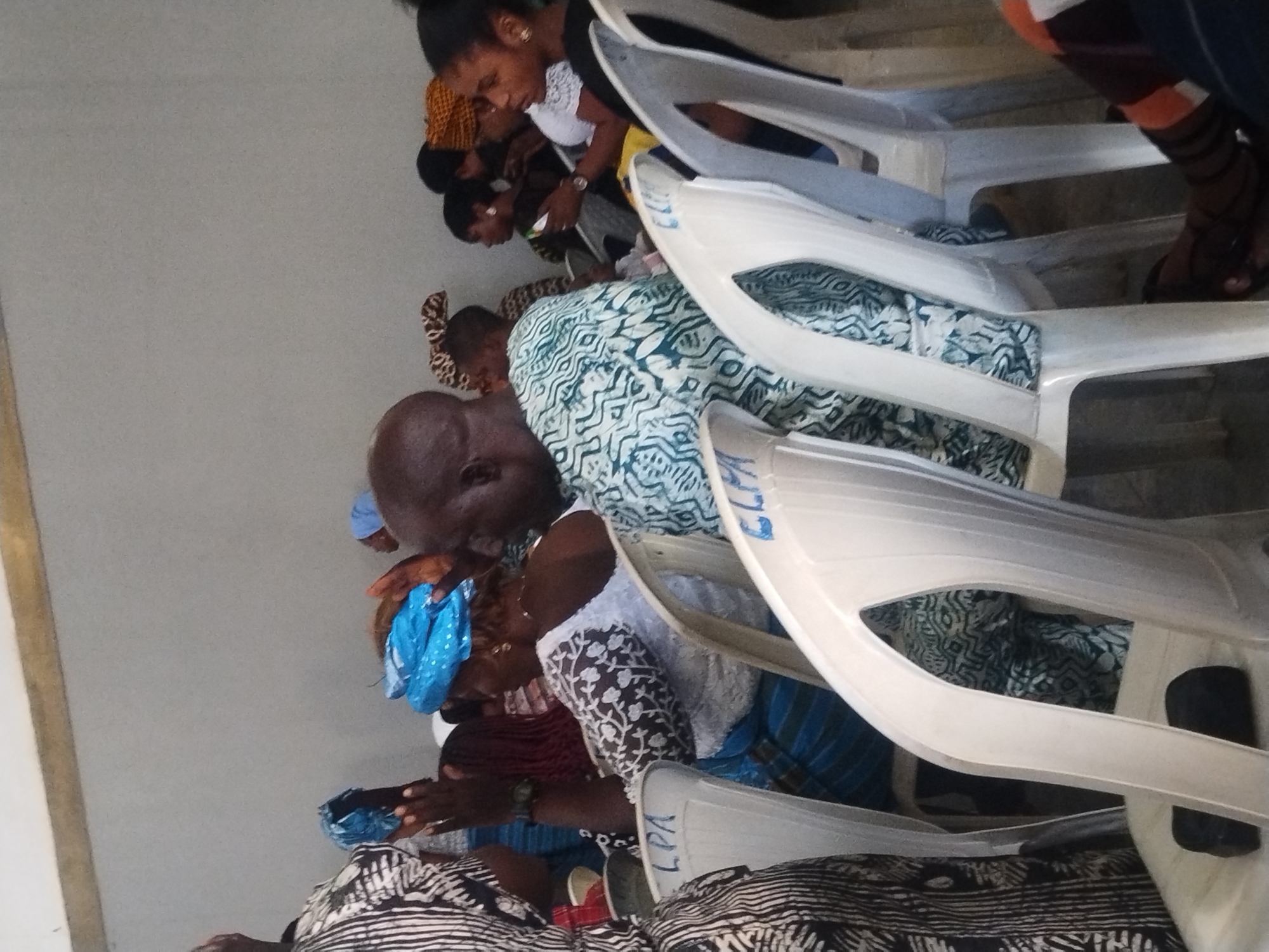 The image shows a group of people sitting in white plastic chairs in what appears to be an indoor space in Kabong, Nigeria. The people are of African descent. Some are looking down, possibly at their laps. They are wearing various patterned clothing, including a blue patterned headscarf, a white and floral print top, and striped fabrics. The chairs are marked with the letters "LPA." The room is plain with light-colored walls.