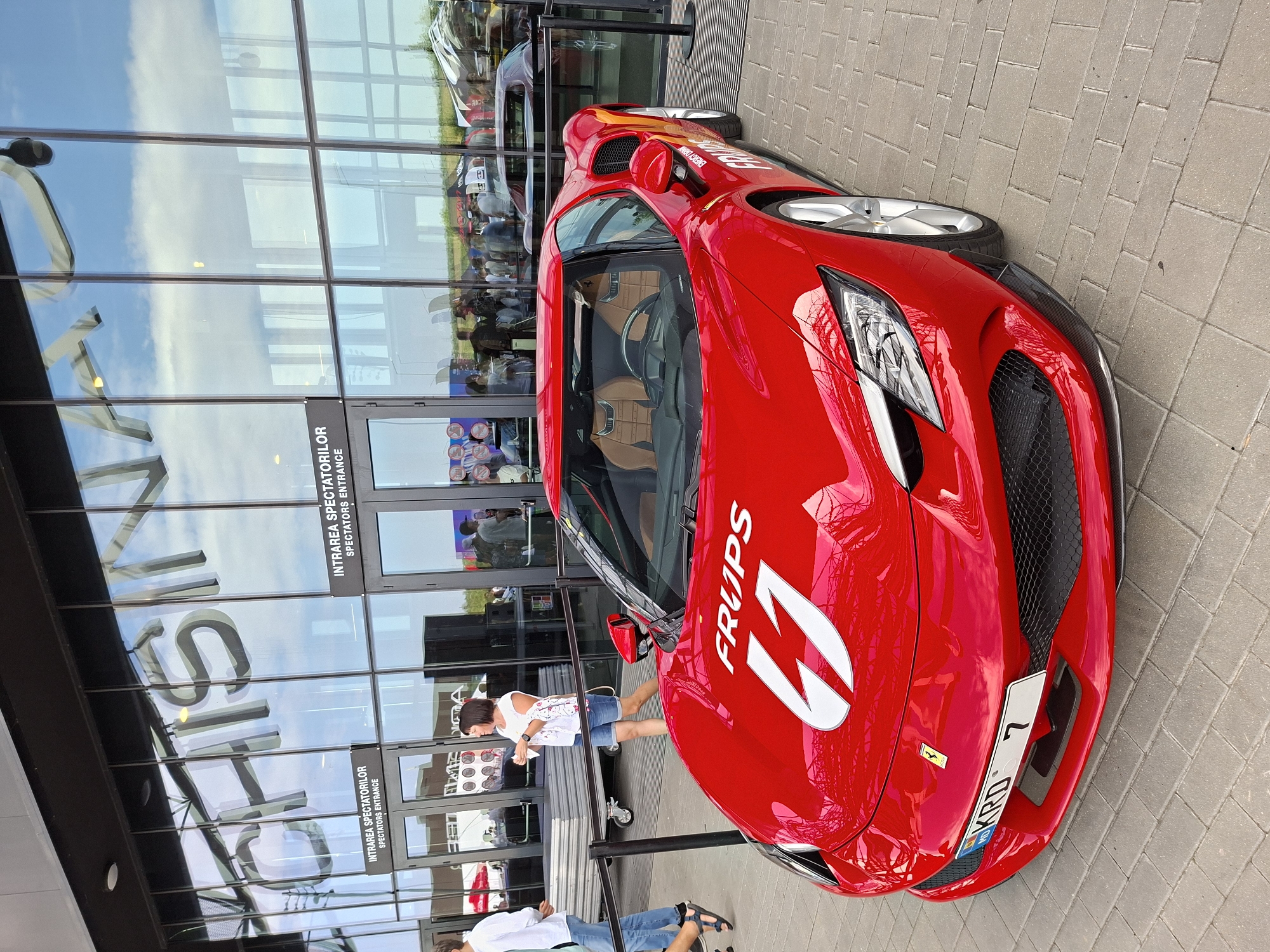 The image shows a red Ferrari parked on a paved surface outside a modern building with a glass facade. The building has large lettering on the glass that appears to spell out "CHISINAU". There are signs above the entrance doors that read "INTRAREA SPECTATORILOR" and "SPECTATORS ENTRANCE". A woman in a white top and denim shorts is standing near the entrance, and other people are visible in the background, some appearing to be spectators. A man in a white shirt and jeans is walking away from the car on the left. The car has "FRUPS" and the number "41" in white decals on its hood, and a license plate that reads "KRD 7". The scene appears to be outdoors, possibly at an event or exhibition in Stăuceni, Moldova, given the building signage.