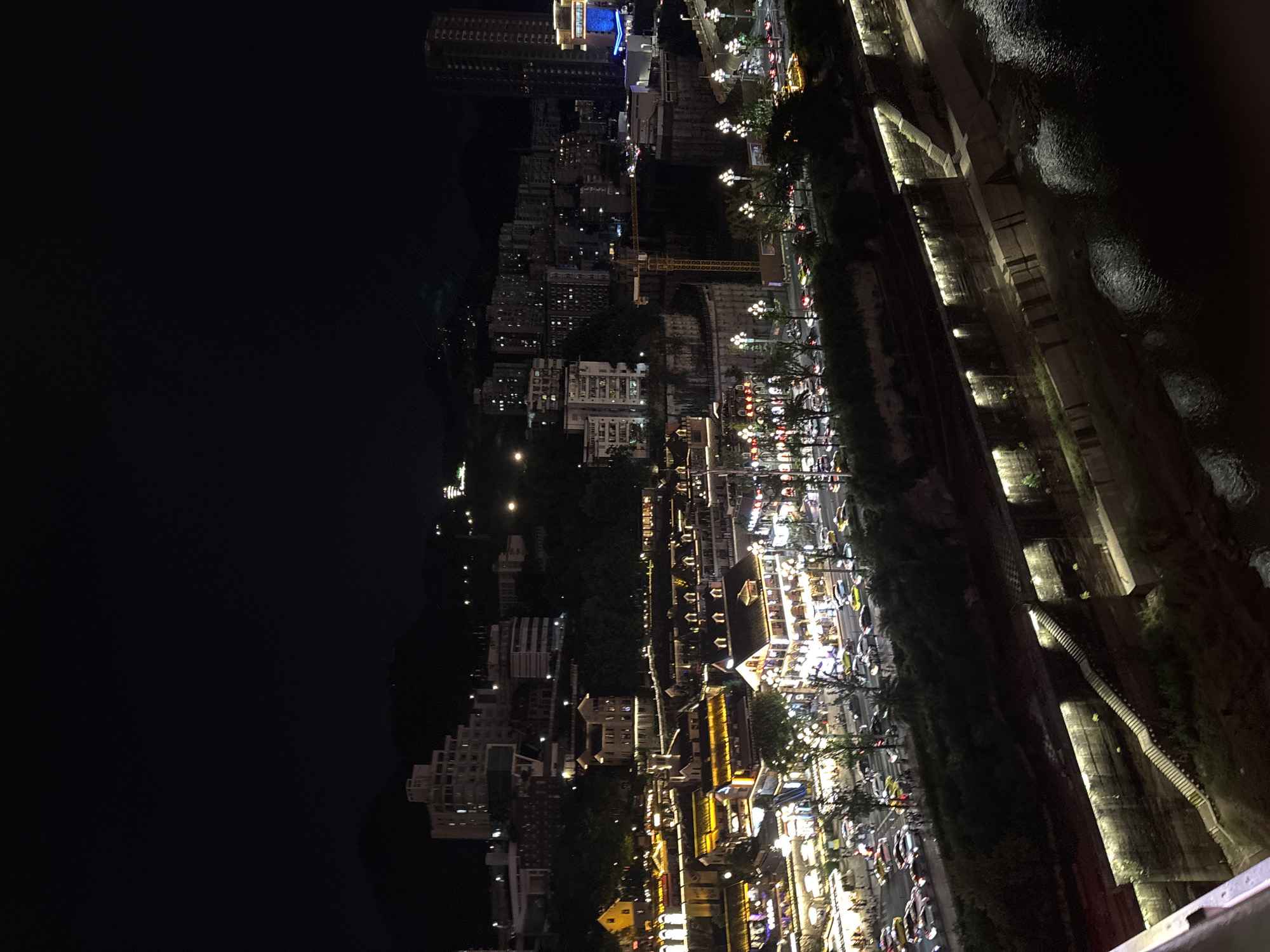 This is a nighttime aerial photograph of a city illuminated by numerous lights. The scene depicts a bustling urban environment with buildings of varying heights, a riverbank, and a body of water. The architecture appears to be a mix of modern high-rise structures and older, more traditional buildings, many of which are adorned with colorful lights, suggesting commercial or residential areas.
Along the riverbank, there's a lively street scene with traffic, indicated by the headlights and taillights of vehicles. Streetlights and the lights from businesses create a vibrant, energetic atmosphere. There are also pedestrian areas with lampposts and trees, adding to the urban landscape. The river itself reflects the lights, creating a shimmering effect on its surface.
The time of day is clearly night, as evidenced by the darkness of the sky and the prevalence of artificial lighting. The weather appears clear, allowing for good visibility of the city lights. The location is hinted at being in Yuzhong District, China, which suggests a potentially scenic riverfront area. The overall mood conveyed by the image is one of vibrancy and activity, a city alive with light and movement after dark. There are no discernible people or specific events taking place, but the scene implies a thriving urban community. No visible text is present in the image.