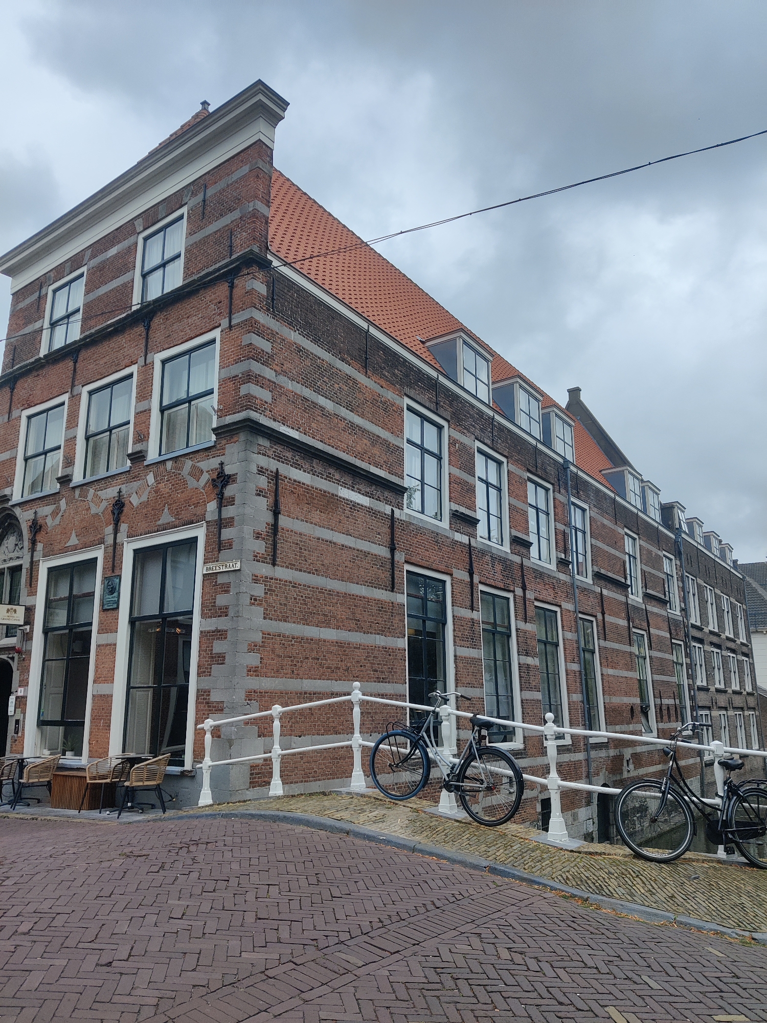 A corner view of a brick building in Delft, Netherlands, shows a street with cobblestone pavement and a white railing along a canal. Two bicycles are parked against the railing. The building has a red tiled roof and multiple windows. A street sign indicates "BREESTRAAT". In the foreground, tables and chairs are set up outside a building, likely a cafe or restaurant. The sky is overcast.