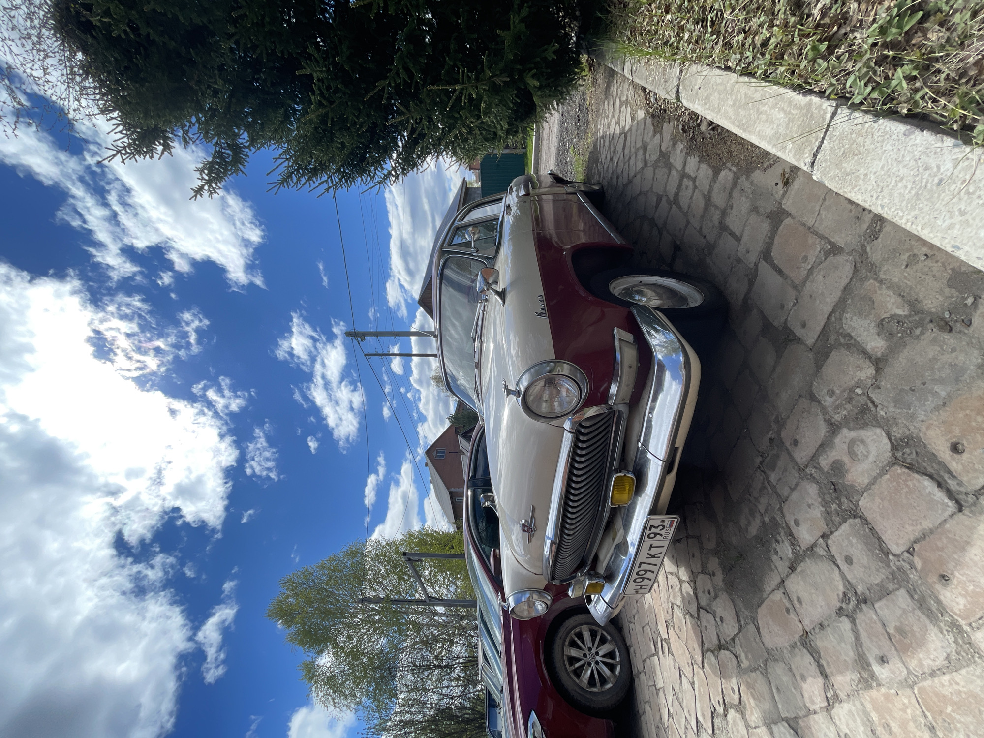 A two-toned maroon and beige GAZ-21 Volga, a vintage Soviet car, is parked on a cobblestone street in Минино, Russia. A portion of another maroon car is visible next to it. The car's license plate is visible, with the code "H997 KT 93 RUS." A sidewalk made of stone slabs runs along the edge of the street. A coniferous tree is located on the left side of the frame. Overhead, a blue sky is scattered with white clouds. Power lines are visible cutting across the sky. A house is partially visible behind the cars.