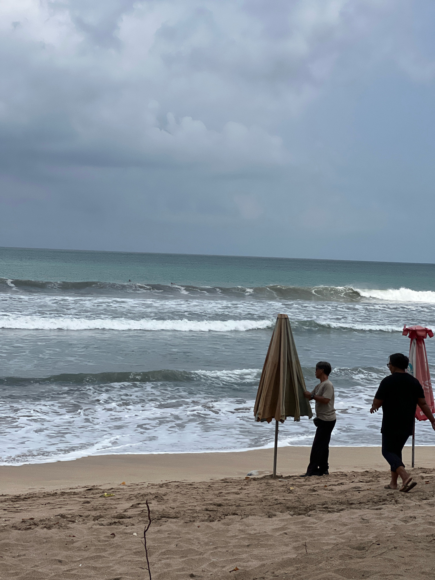 The image depicts a beach scene in Kuta, Indonesia, under a cloudy sky. Two individuals are present on the sandy shore. One person, wearing a light-colored t-shirt and dark pants, stands near a closed beach umbrella, adjusting it. Another person, in a black t-shirt and dark pants, walks away from the viewer towards the right side of the frame, also near a closed beach umbrella. The ocean is visible in the background, with moderate waves breaking and foam washing ashore. Several surfers can be seen in the distance on the water. The overall atmosphere is overcast, suggesting potential rain or a stormy day. The sand is a light brown color and appears somewhat damp near the water's edge.