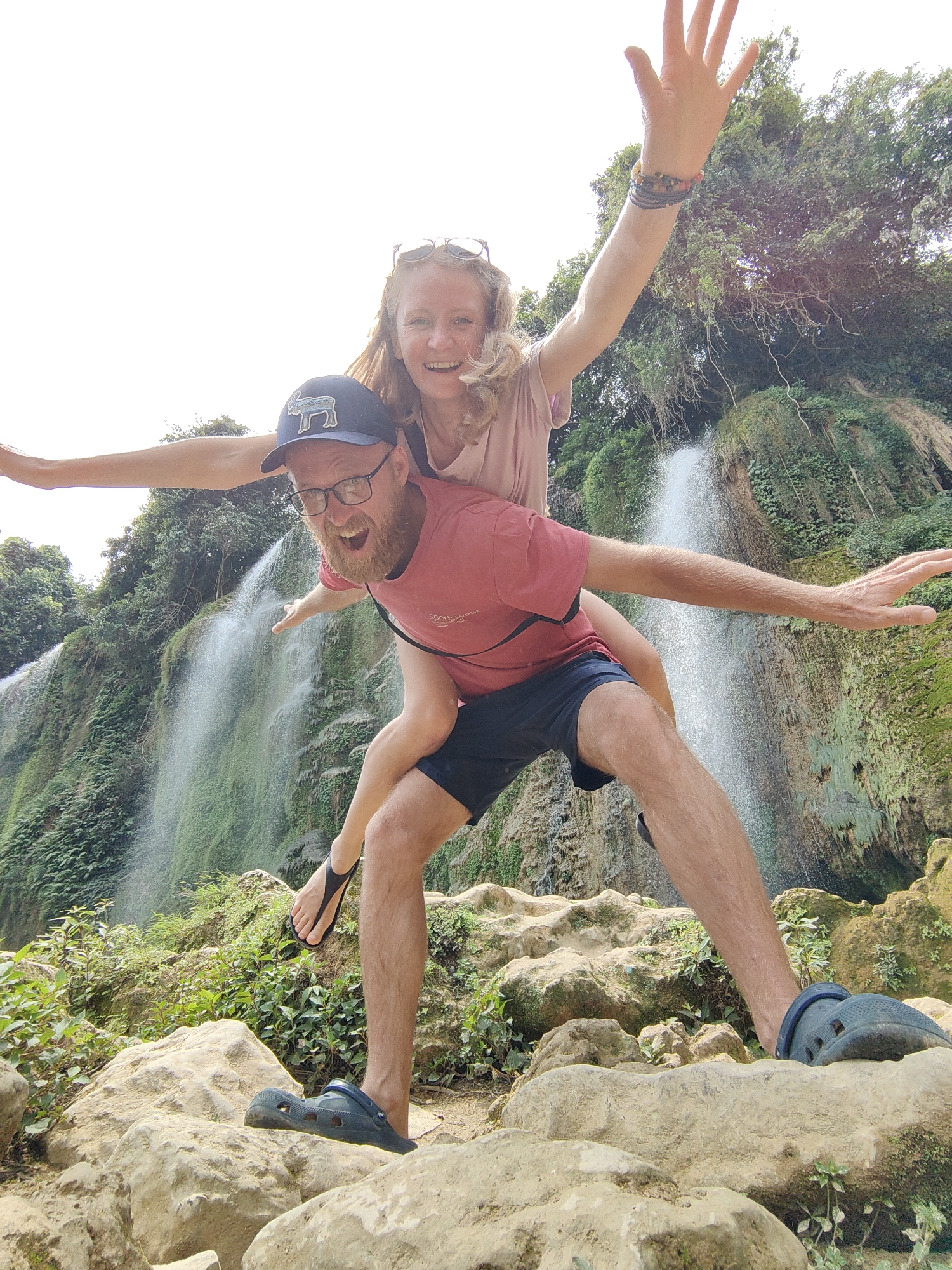 The photograph shows a couple posing in front of a waterfall. The man is giving the woman a piggyback ride. He is wearing a red t-shirt, blue shorts, a dark blue baseball cap with a logo of an animal, glasses, and blue Crocs on his feet. The woman is wearing a light pink t-shirt and has her arms outstretched. She also has sunglasses on top of her head and a bracelet on her right wrist. They are standing on rocks covered in greenery. The waterfall is in the background, surrounded by lush green vegetation. The location appears to be within Daxin County, China, given the landscape.