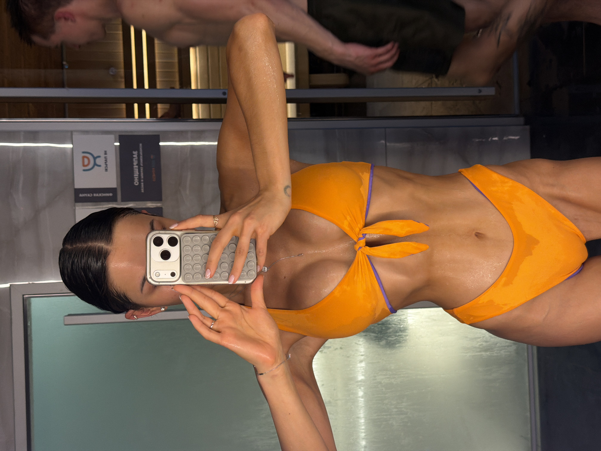 The image, presented rotated 90 degrees clockwise, depicts a woman taking a mirror selfie in an indoor spa or pool environment. The primary subject is a young woman with dark, slicked-back hair, wearing a vibrant orange bikini with purple trim. Her skin is visibly wet and glistening, indicating recent exposure to water. She holds a light-colored smartphone with a distinct textured case, angled to capture her reflection. Notable personal details include several rings on her fingers, a delicate silver necklace, a matching bracelet, and a small crescent moon tattoo on her inner left forearm.
Reflected in the background behind her is a partially visible man, also appearing wet and possibly shirtless or in swim attire, moving within the same facility. The setting features elements typical of a modern spa or aquatic center, including horizontal wooden paneling, reflective glass or mirror surfaces, and dark wall tiles.
Two signs are prominently reflected. The left sign displays a "DC" logo along with Cyrillic text, including "ОЛИМПИЙСКИЙ" (Olimpiyskiy), suggesting "Olympic," and "АНУАЛ РАЖННО" (Anual Razhno), although this latter text is somewhat obscured or distorted by reflection. The right sign clearly states "ЗАПРЕЩЕНО" (Zapreshcheno), which translates to "FORBIDDEN" or "PROHIBITED," and "СПОРТ" (Sport) below it. These Russian texts strongly confirm the location context of Moscow, Russia. The image captures a candid moment of leisure and self-documentation. The indoor lighting suggests no specific time of day but appears bright.