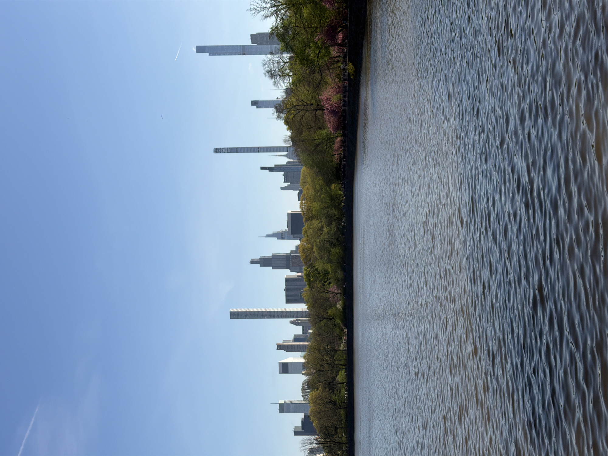 The image captures a cityscape view alongside a body of water, likely a reservoir or pond in Central Park, New York City. The perspective is oriented vertically, with the sky dominating the left side of the frame. The skyline features an array of skyscrapers, some obscured by trees, indicating a park setting. The right side is occupied by the water, its surface textured by ripples and reflections. The day appears clear, with a blue sky. The composition suggests a blend of urban development and natural landscape within New York City.