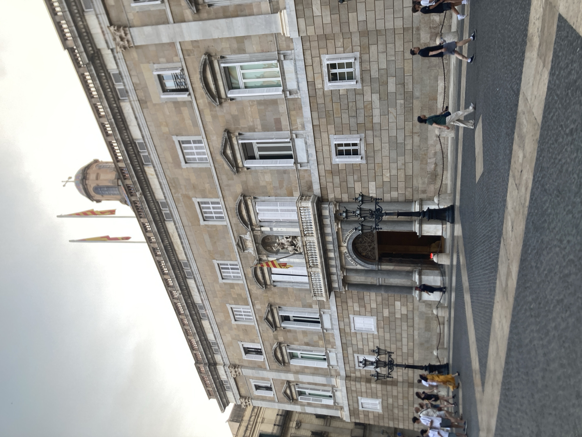 The image shows the Palau de la Generalitat de Catalunya in Barcelona, Spain. The building is a large, stone structure with a classical facade, featuring numerous windows and decorative elements. Two flags are flying from the rooftop, and a dome with a cross is visible in the background. In the foreground, a group of people are walking on a cobblestone plaza. Some are tourists, taking in the sights, while others appear to be locals going about their day. The sky is overcast, suggesting a cloudy day. The lighting is soft, with no strong shadows, indicating it is likely late afternoon or early morning. The overall impression is one of a grand historical building in a public space, with people interacting with their surroundings.
