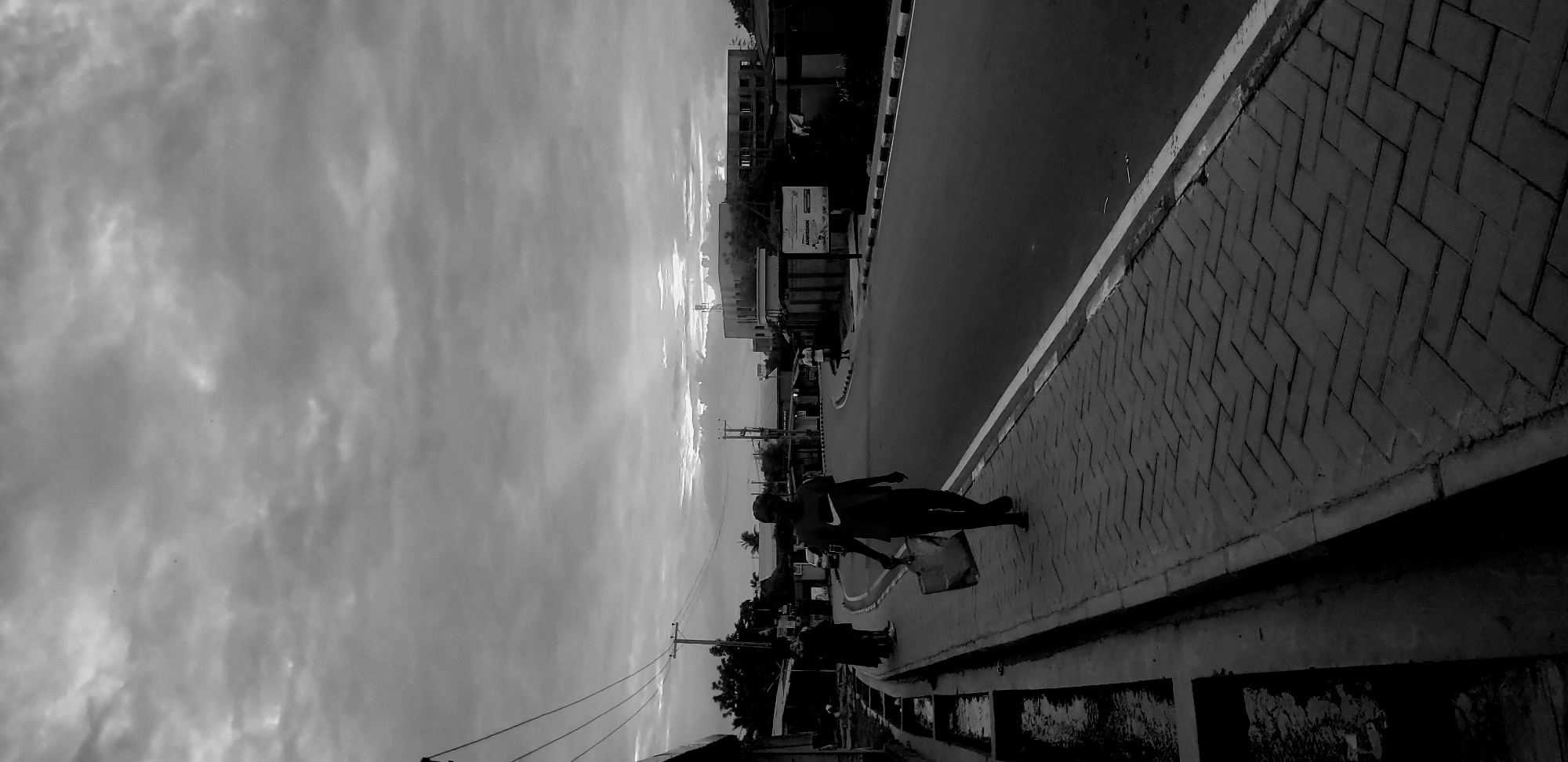The black and white image displays a street scene in Narayi, Nigeria, under an overcast sky with diffused light. Heavy grey clouds are visible across the upper frame, with brighter areas indicating obscured light sources. In the foreground, a brick-paved sidewalk with a herringbone pattern runs along the right side, adjacent to a concrete drainage channel containing stagnant water on the far left. A male individual, wearing a dark t-shirt with a white Nike swoosh logo and dark trousers, walks on the sidewalk towards the viewer, carrying a light-colored bag in his left hand.
The asphalt road, delineated by a continuous white line on the right, curves gently to the right into the mid-ground. Another male figure, dressed in a lighter shirt, stands near the curb on the right side of the road in the mid-ground. Along the left side of the street, multiple utility poles with complex arrangements of overhead electrical wires are visible, fronting a series of low-rise buildings, some appearing residential or commercial.
On the right side of the mid-ground, a rectangular sign displaying the word "ADMISSION" is affixed to a perimeter wall. Behind this wall, multi-story buildings are present; one structure appears to be under construction or partially derelict, while an adjacent building features a prominent antenna or mast on its roof. In the distant background, a faint elevated structure, possibly a bridge, spans across the scene. Indistinct figures are also visible on the far left sidewalk.