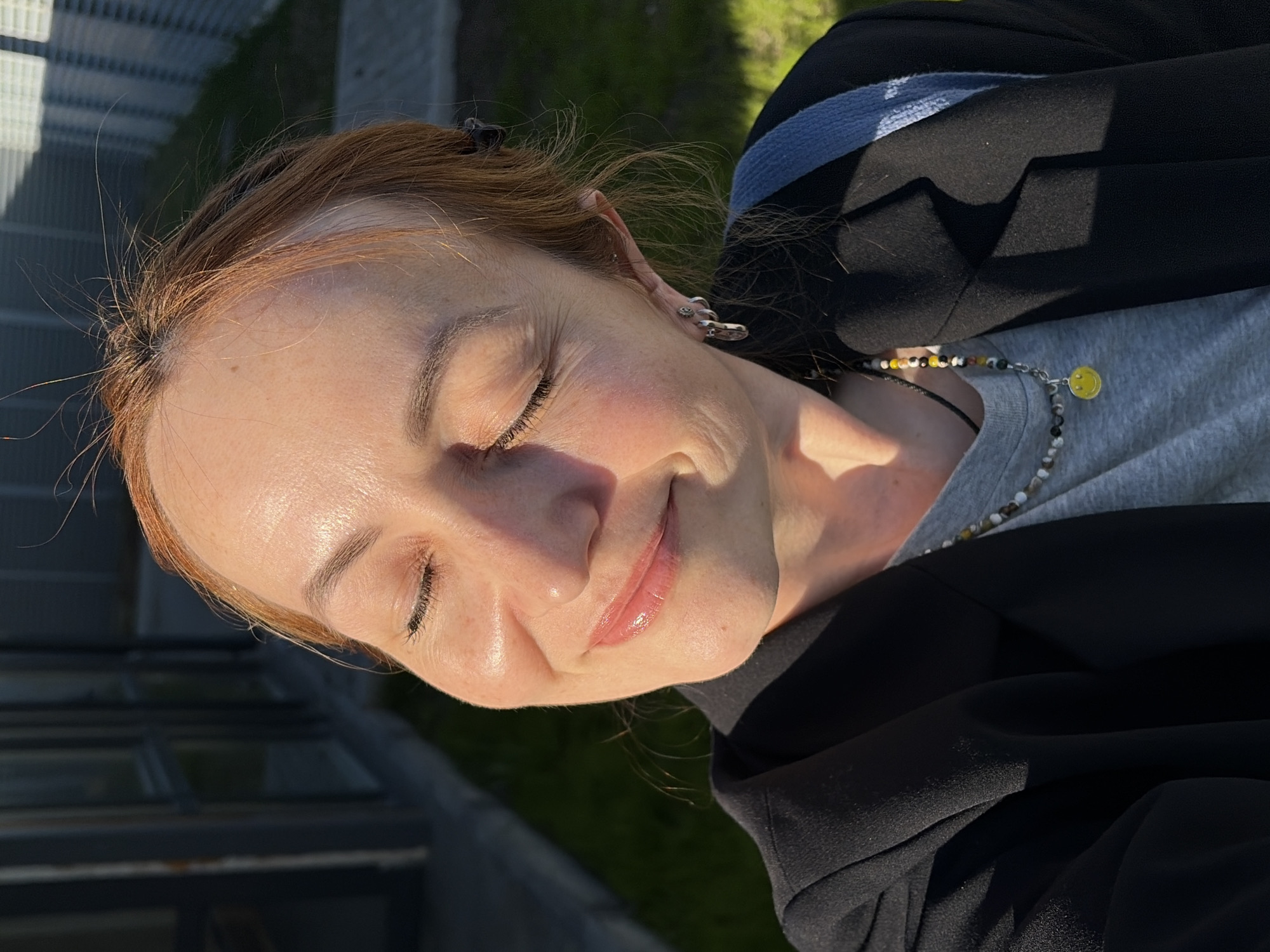 A fair-skinned woman with reddish-blonde hair is pictured in a medium shot taken outdoors during daylight. The woman's eyes are closed, and she has a slight smile. She wears a grey t-shirt, a black jacket, and a beaded necklace with a yellow smiley face charm. Her left ear is visible, adorned with earrings. A building's glass facade and some greenery appear in the background. The location is Chaikovsky, Russia.