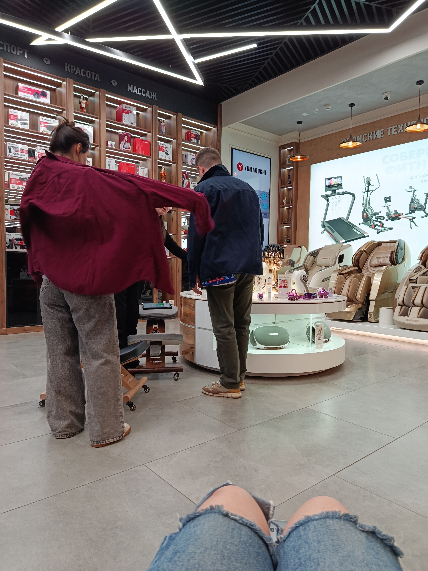 The photograph appears to have been taken in a retail store in Moscow, Russia, judging by the Cyrillic lettering on the signs and displays. The store seems to specialize in massage and fitness equipment, featuring items such as massage chairs, treadmills, and other health and beauty products.
Several people are visible in the shot. In the foreground, a person is wearing distressed denim shorts and appears to be sitting down, with their knees visible. Further back, two people are interacting with a store employee near a display of products. One of the individuals is wearing a maroon shirt and gray pants, while the other is wearing a navy blue jacket and olive green pants.
The store has a modern design with bright lighting, geometric ceiling patterns, and wooden shelving units. The products are neatly arranged on the shelves and on a circular display table. The overall impression is that of a clean, organized, and well-lit retail environment.