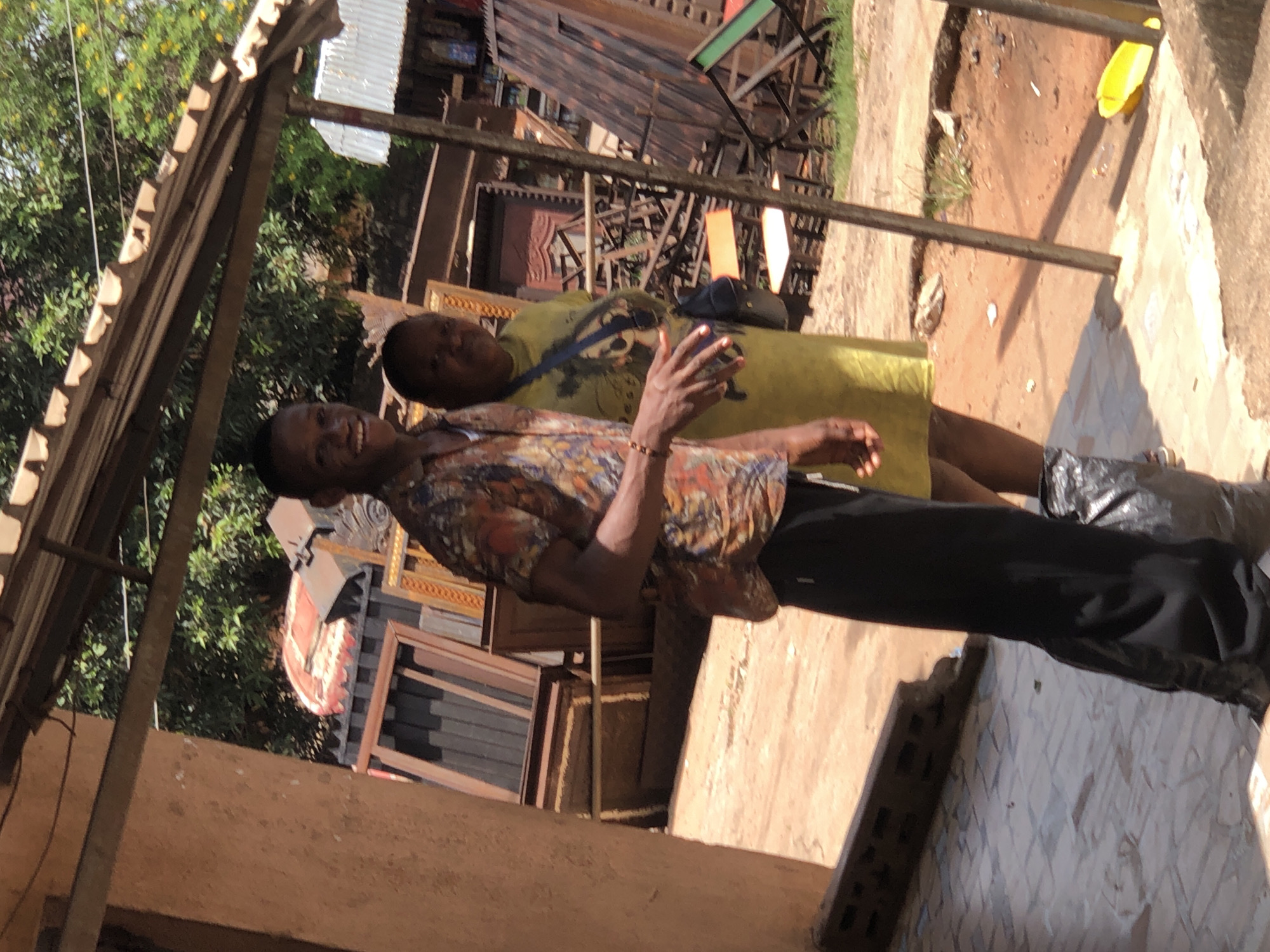 Outside in Onitsha, Nigeria, two people are visible. One individual is standing and smiling, wearing a colorful short-sleeved shirt and dark trousers. The second person is standing behind the first, wearing a light yellow garment and a bag slung across their shoulder. A collection of wooden frames and furniture is stacked in the background. The setting appears to be near a building or shop with a corrugated metal roof.