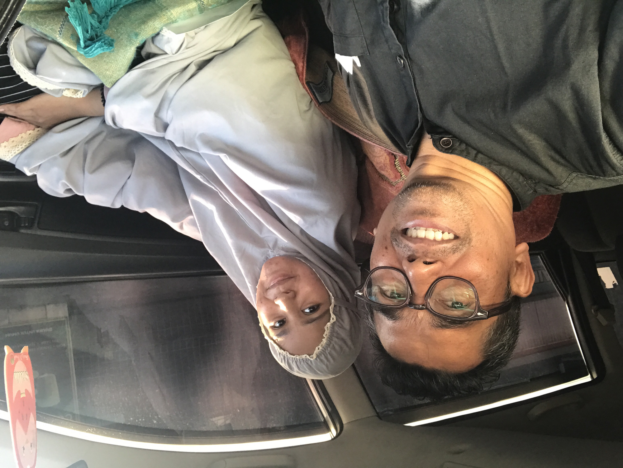Two people are posing for a selfie inside a car. The woman on the left wears a gray headscarf. The man on the right has glasses. The car is moving through the city of Lhokseumawe, Indonesia. The interior of the vehicle contains various objects, including clothing and an air freshener. The car's sunroof is visible in the upper right corner.