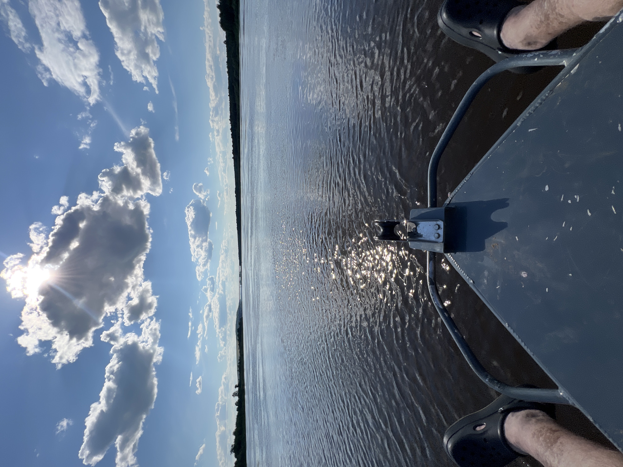 A first-person perspective shot captures the view from the bow of a boat moving across a wide, dark river. The sun, partially obscured by dramatic, fluffy white clouds, shines brightly, creating a shimmering reflection on the water's surface. The riverbanks are lined with dense, dark green trees, and in the distance, a low, undulating hill is visible. The foreground shows the grey, metallic bow of the boat, with a black winch mechanism mounted in the center. On either side of the bow, a pair of black Crocs is visible, with the wearer's bare legs extending from beneath the frame. The sky is a vibrant blue, with scattered white clouds and a bright sun near the top center. The overall impression is one of serene travel on a sunny day, with the user's feet casually resting as the boat navigates the waterway. The location is the city of Sosnovka, Russia.
