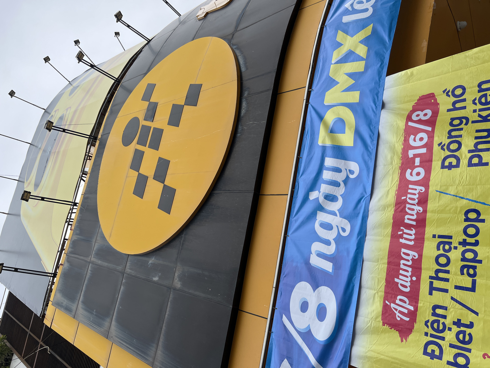 The image captures a close-up, low-angle view of a building facade in Nha Trang, Vietnam. The building features a large, curved yellow circular logo with a black, pixelated pattern in the center, set against a dark grey tiled surface. Below this, a bright blue banner displays white text, partially visible, reading "8 ngày DMX le". Further down, a vibrant yellow banner is predominantly visible, with a red rectangular section at the top containing white text that reads "Áp dụng từ ngày 6-16/8". Below this red section, blue text is visible, with "Điện Thoại" and "blet / Laptop" appearing on the left, and "Đồng hồ" and "Phu kiện" on the right. The banners suggest a commercial promotion, likely for electronics or mobile devices. The overall lighting suggests an overcast day.