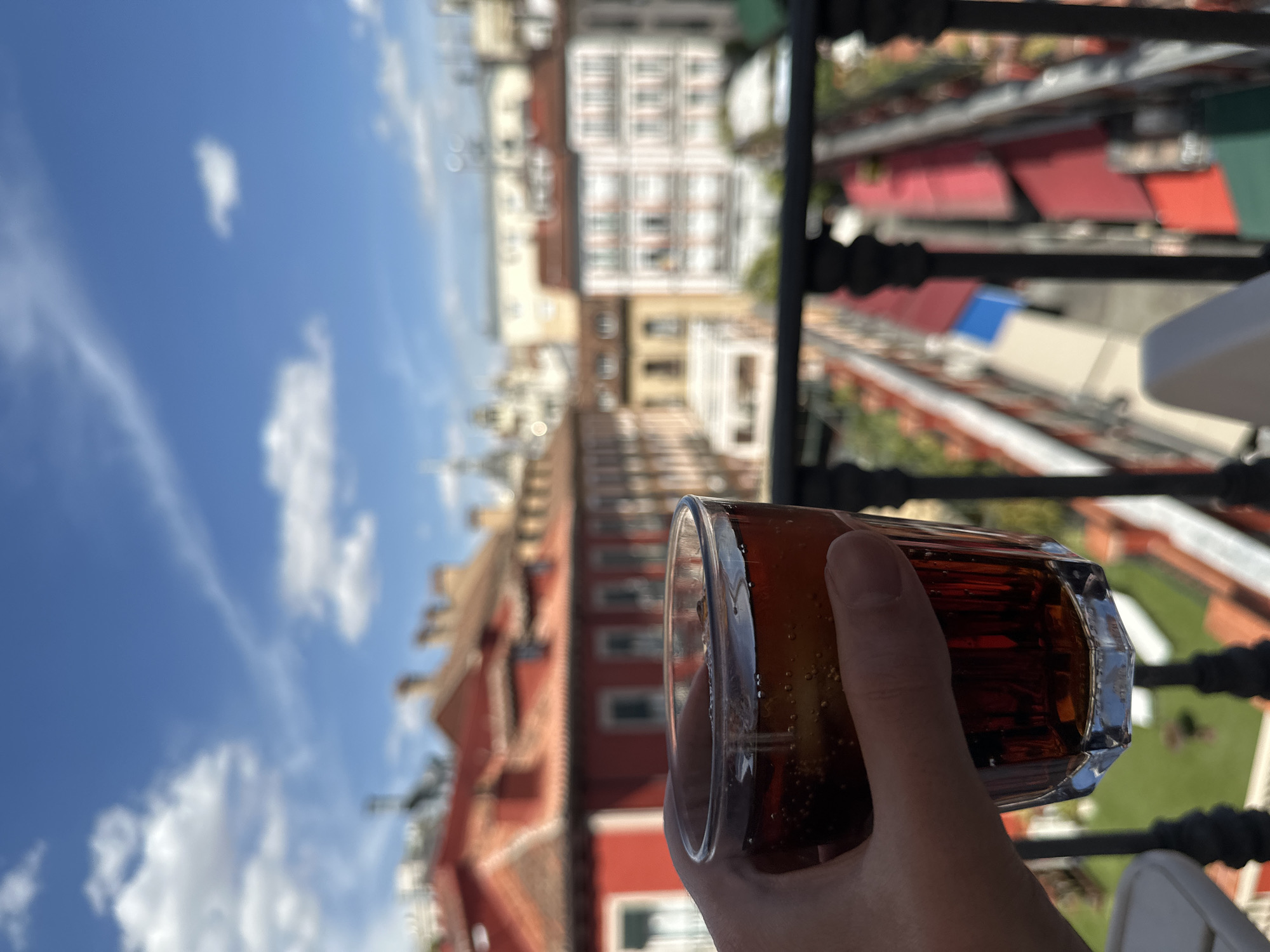 A close-up view shows a lightly tanned right hand holding a faceted glass containing a dark, carbonated beverage with ice cubes and visible bubbles. The glass is positioned in the lower-left foreground, slightly angled. Directly in front of the glass and below it, a black wrought-iron balcony railing runs horizontally. The background, out of focus, reveals a vibrant city scene typical of Madrid, Spain. Prominently featured are multiple multi-story buildings with reddish-brown facades and distinctive terracotta tiled roofs, some exhibiting chimneys. Further into the distance, lighter-colored buildings with balconies are discernible. Below these structures, a bustling street or pedestrian area is suggested by numerous colorful awnings, predominantly red, with some blue and green, indicating outdoor cafes, restaurants, or market stalls. The upper portion of the image is dominated by a bright blue sky with scattered white cumulus clouds, indicating a sunny daytime scene. No other people or direct interactions are visible. The overall environment suggests an urban setting, likely a balcony or rooftop terrace overlooking a historic district in central Madrid.