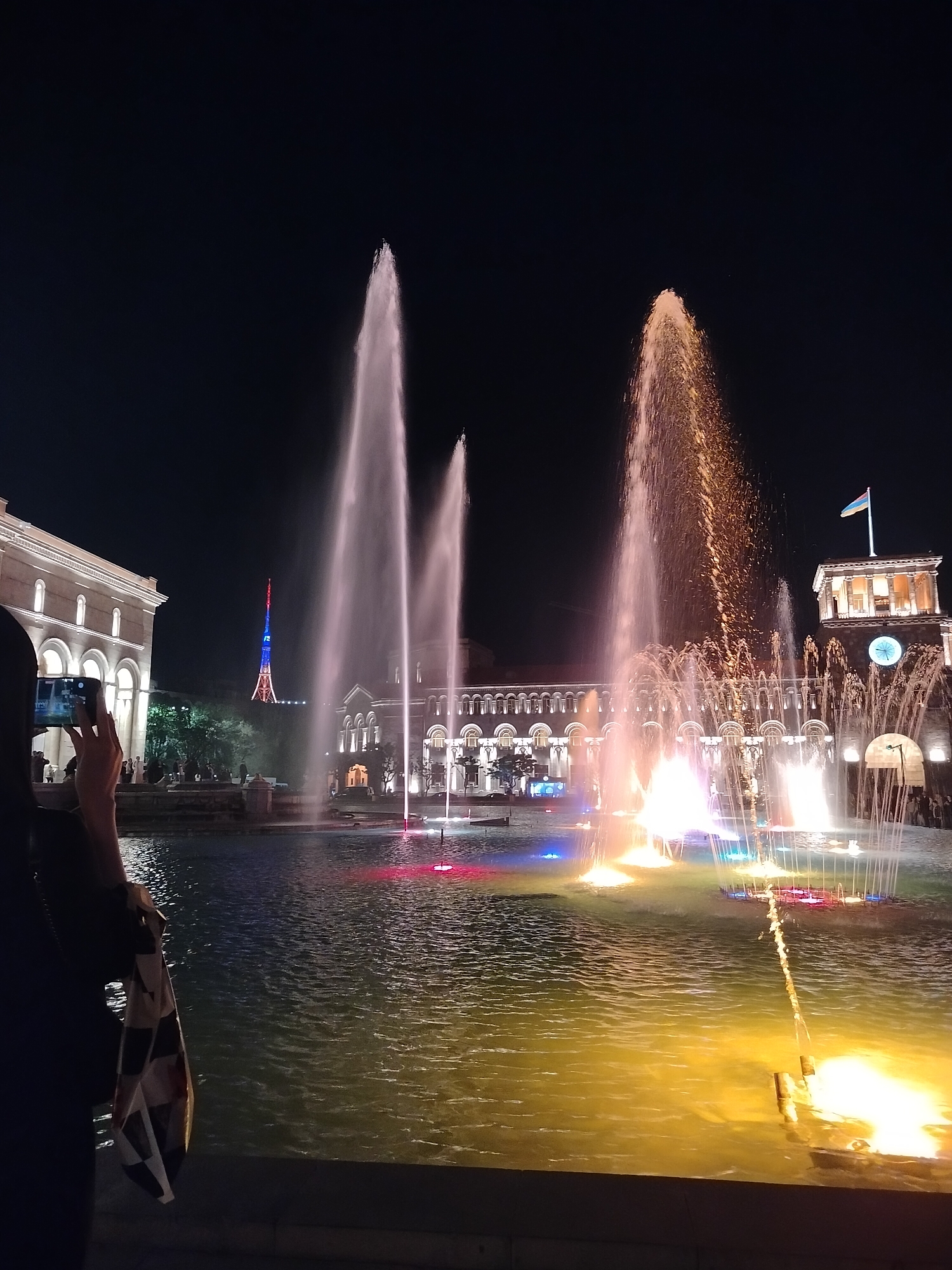 The image depicts a night scene at Republic Square in Yerevan, Armenia. In the foreground is a large fountain with multiple water jets illuminated by colored lights, creating red, blue, yellow, and orange hues in the water. To the left, a person is holding a smartphone to take a photo. Behind the fountain is a grand building with arched windows, lit up with white lights. A clock tower with a flag on top is visible on the right side of the building. In the distance, a tower structure illuminated with red, blue, and white lights can be seen. The sky is dark.