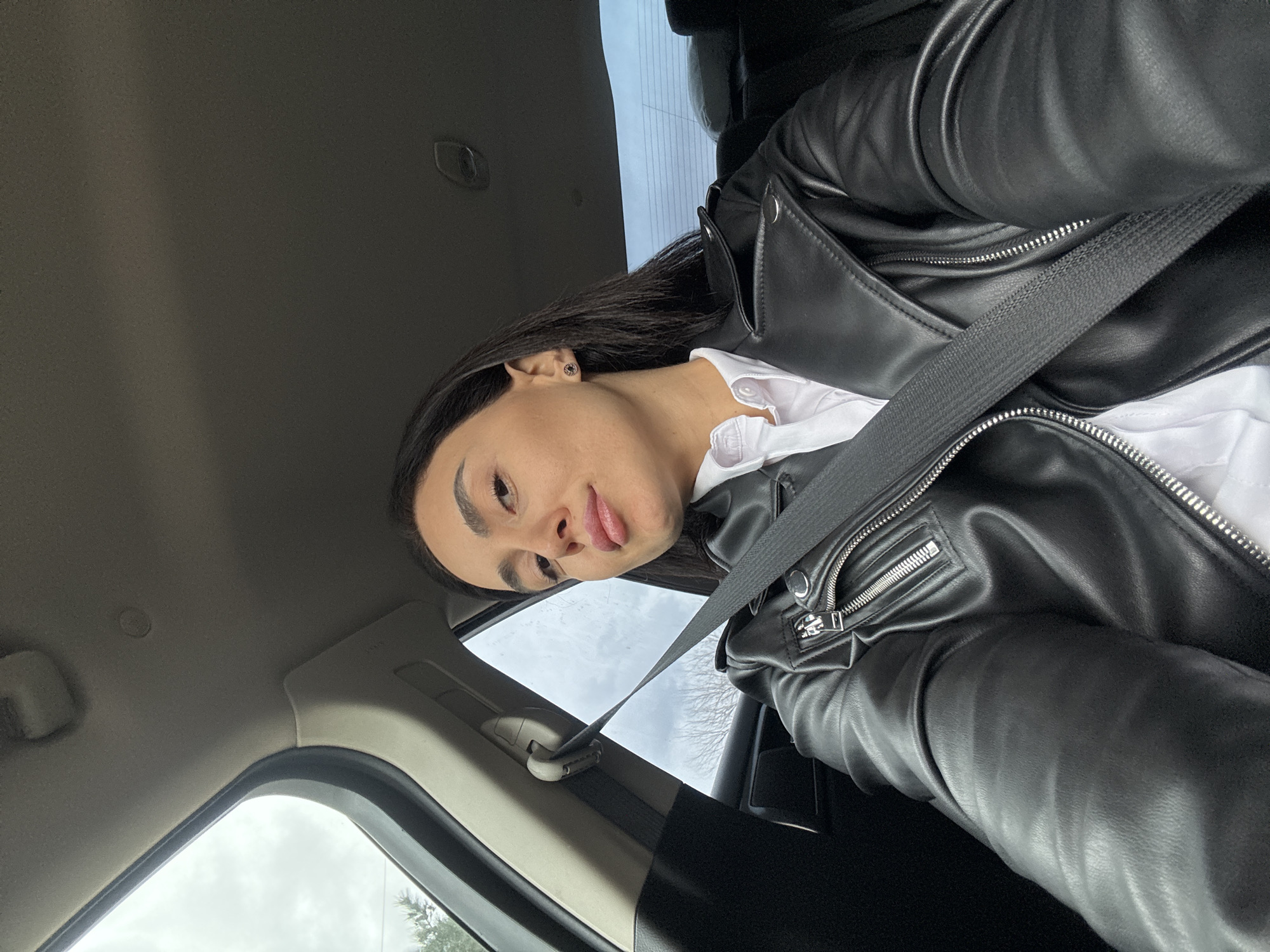 A person is seated in a vehicle, wearing a black leather jacket over a white collared shirt and a seatbelt. The interior of the car is visible, including parts of the door and the roof. Through the car window, bare trees are visible against a cloudy sky. The location cannot be ascertained as Tbilisi, Georgia.