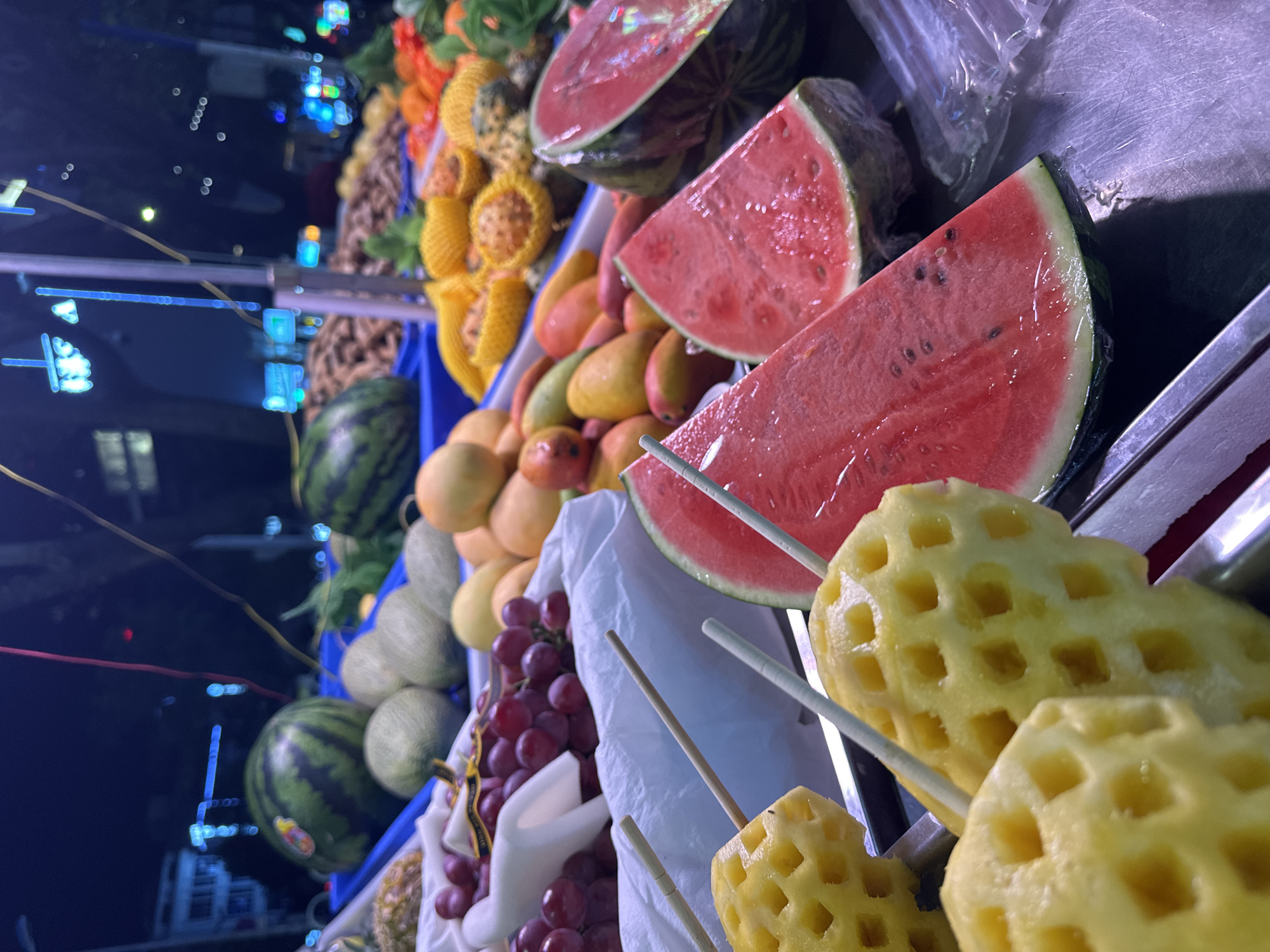 This is a nighttime, eye-level, close-up shot of a fruit stand in Kunming, China. The scene is dominated by a variety of fruits, with a focus on sliced watermelons and pineapple chunks on skewers in the foreground. In the background, other fruits like grapes, mangoes, and what appear to be cantaloupes are visible, along with some dried goods. The stand is illuminated by artificial lights, casting a colorful glow on the produce. Beyond the fruit stand, a blurred cityscape with glowing lights suggests an urban environment at night. The overall impression is one of a vibrant and bustling street market.
