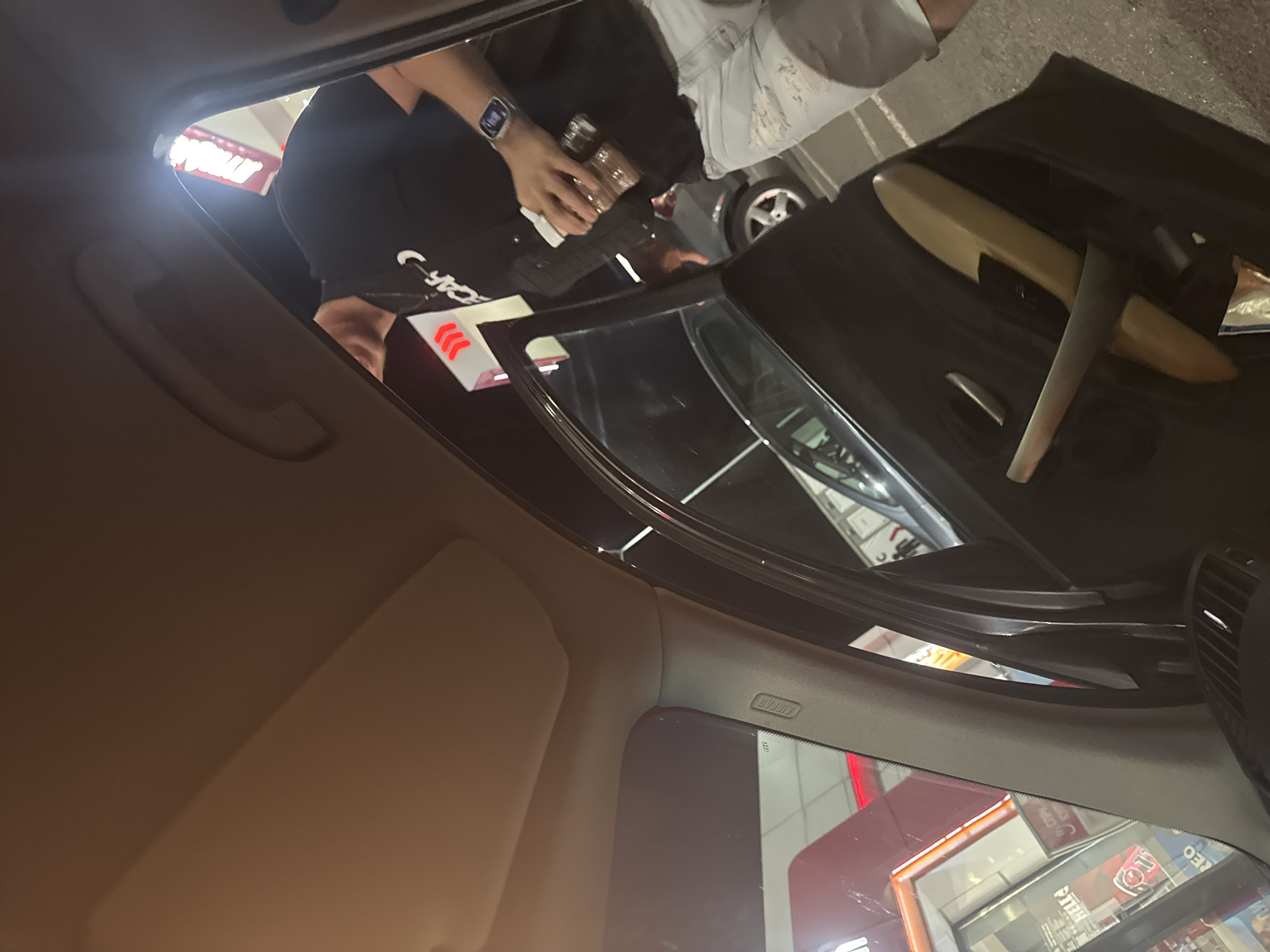 The image depicts the interior of a car, viewed from the driver's perspective. Outside the car, an individual wearing a black shirt and a watch is holding a cup. The car appears to be at a drive-through, where a box or container with red and white stripes is visible. In the reflection in the rearview mirror, a building with a red sign can be seen. Given the location constraints, there is no information to verify if the scene occurred in Vodenichane, Bulgaria.