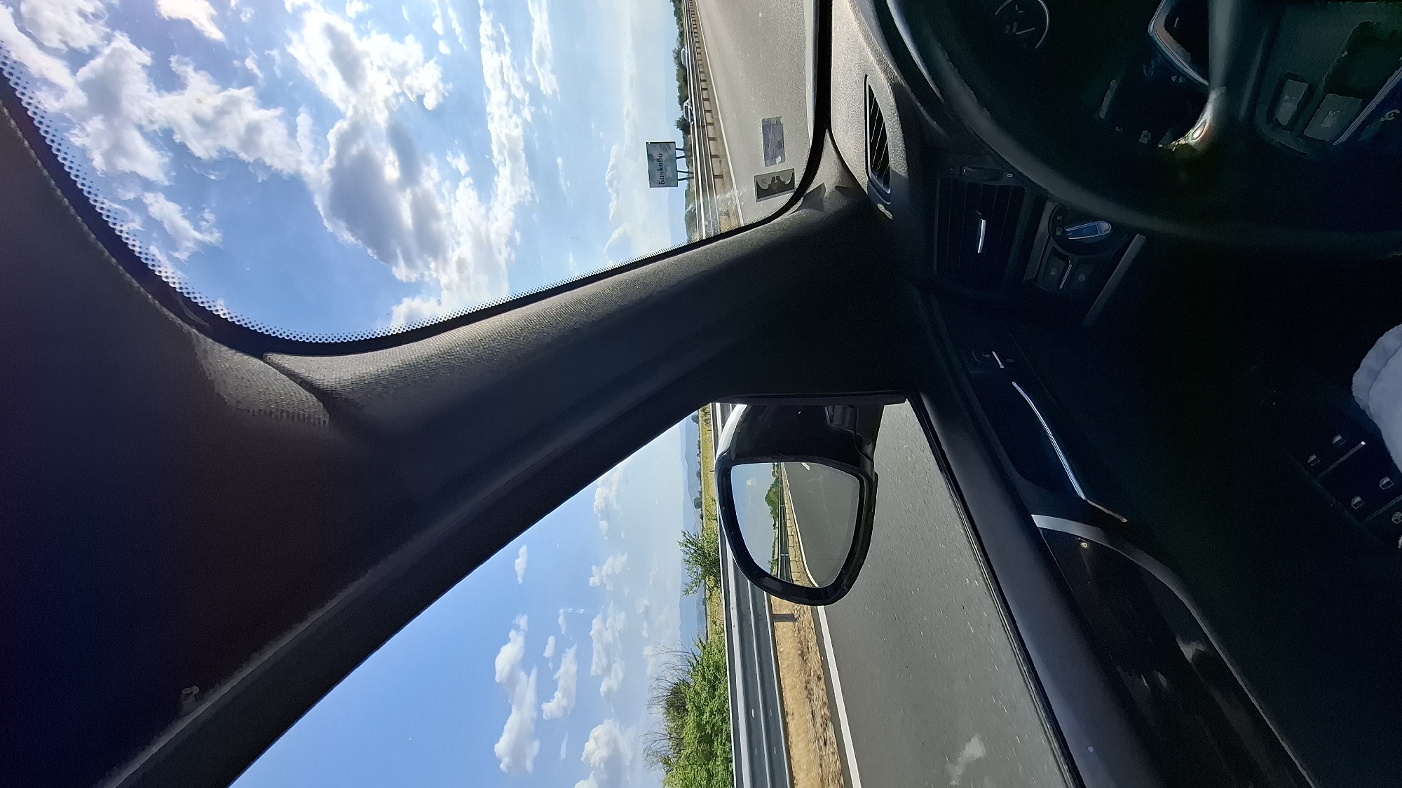 A first-person perspective captures the view from inside a car traveling on a highway under a bright, cloudy sky. The car's interior, including the dashboard, steering wheel, and a portion of the driver's side door, is visible in the foreground. A side mirror reflects the highway and surrounding landscape. Outside the car, the highway stretches ahead, bordered by a guardrail and vegetation. In the distance, a billboard with Cyrillic text, which reads "Бачкову" (Bachkovu), is visible, indicating the location is near Malo Konare, Bulgaria. The scene is a snapshot of a road trip, emphasizing the journey and the external environment.