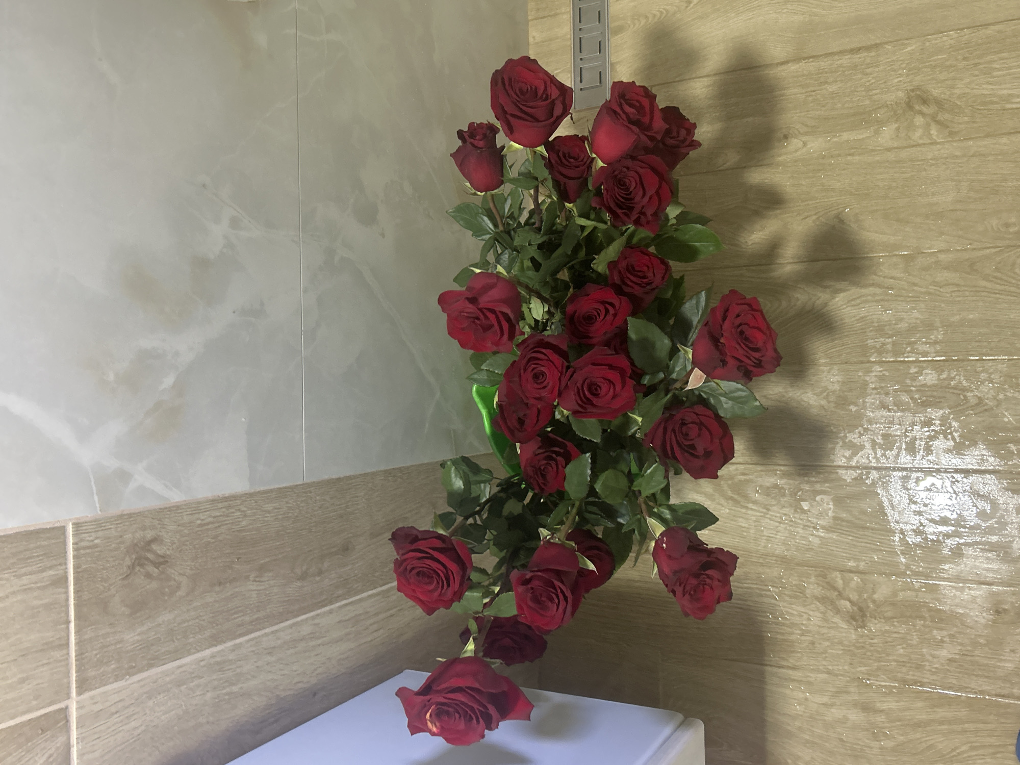 A vertical, eye-level shot shows a bouquet of red roses arranged in a green vase, placed on a white surface next to a wall. The wall to the left has a light gray marble-like pattern, while the wall to the right has a horizontal light brown wood grain pattern. One rose has fallen out of the bouquet and rests on the white surface. The bouquet appears to be indoors. There are no people visible. There is no information in the image to specifically identify it as being in the city of Zlatograd, Bulgaria.