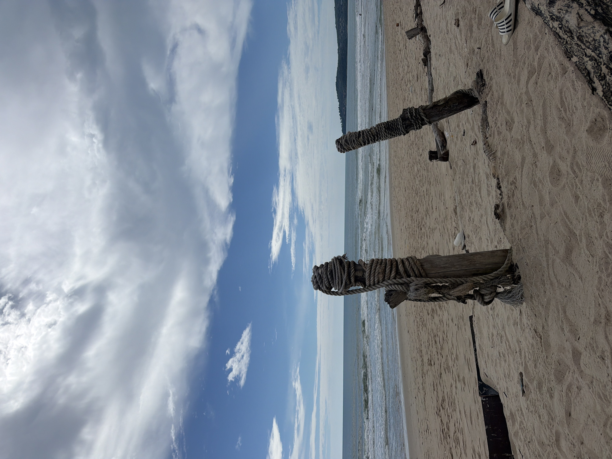 This image depicts a sandy beach scene in Choeng Thale, Thailand, under a partially cloudy sky. In the foreground, two weathered wooden posts are present, each wrapped with thick rope. The post on the left is taller and more substantial, with multiple strands of rope tightly coiled around its upper section and hanging down. The post on the right is shorter and thinner, also wrapped with rope. The beach itself is composed of light-colored sand, with visible footprints and scattered debris including what appears to be a piece of driftwood. In the middle ground, the ocean stretches towards the horizon, with gentle waves rolling onto the shore. The sky is a mix of blue sky and large, scattered white clouds with grey undertones. In the distance, a faint green, tree-covered landmass is visible along the coastline. On the far right of the image, a pair of white sandals with black stripes are placed on the sand. There are no discernible people actively engaged in any actions within the immediate frame, though a distant figure can be faintly seen in the water.