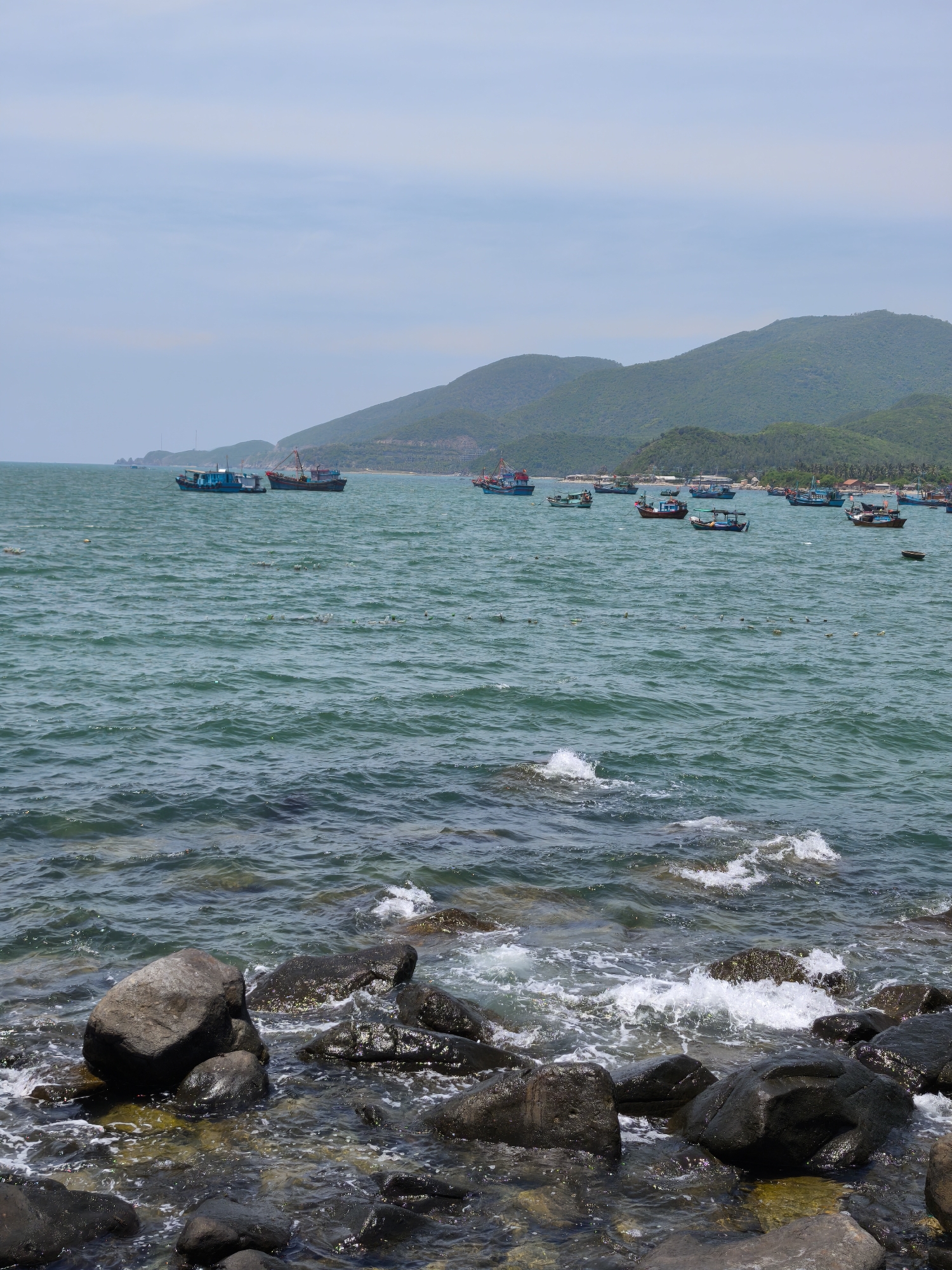 This image depicts a coastal scene in Vĩnh Lương, Vietnam. The foreground is dominated by dark, wet rocks and the incoming waves of a turquoise sea, creating a natural shoreline. In the mid-ground, a multitude of fishing boats are scattered across the water, their hulls varied in color, with some appearing to be anchored or moving slowly. The boats are of different sizes and designs, some with prominent masts and rigging. The water is choppy, with visible ripples and small waves crashing against the rocks in the foreground. In the background, a range of lush green, tree-covered hills rise from the coastline, forming a mountainous backdrop. The sky above is a pale blue with scattered, wispy clouds, suggesting fair weather. The overall impression is of a working fishing harbor or a scenic bay with active maritime elements. There are no people clearly visible in the image.