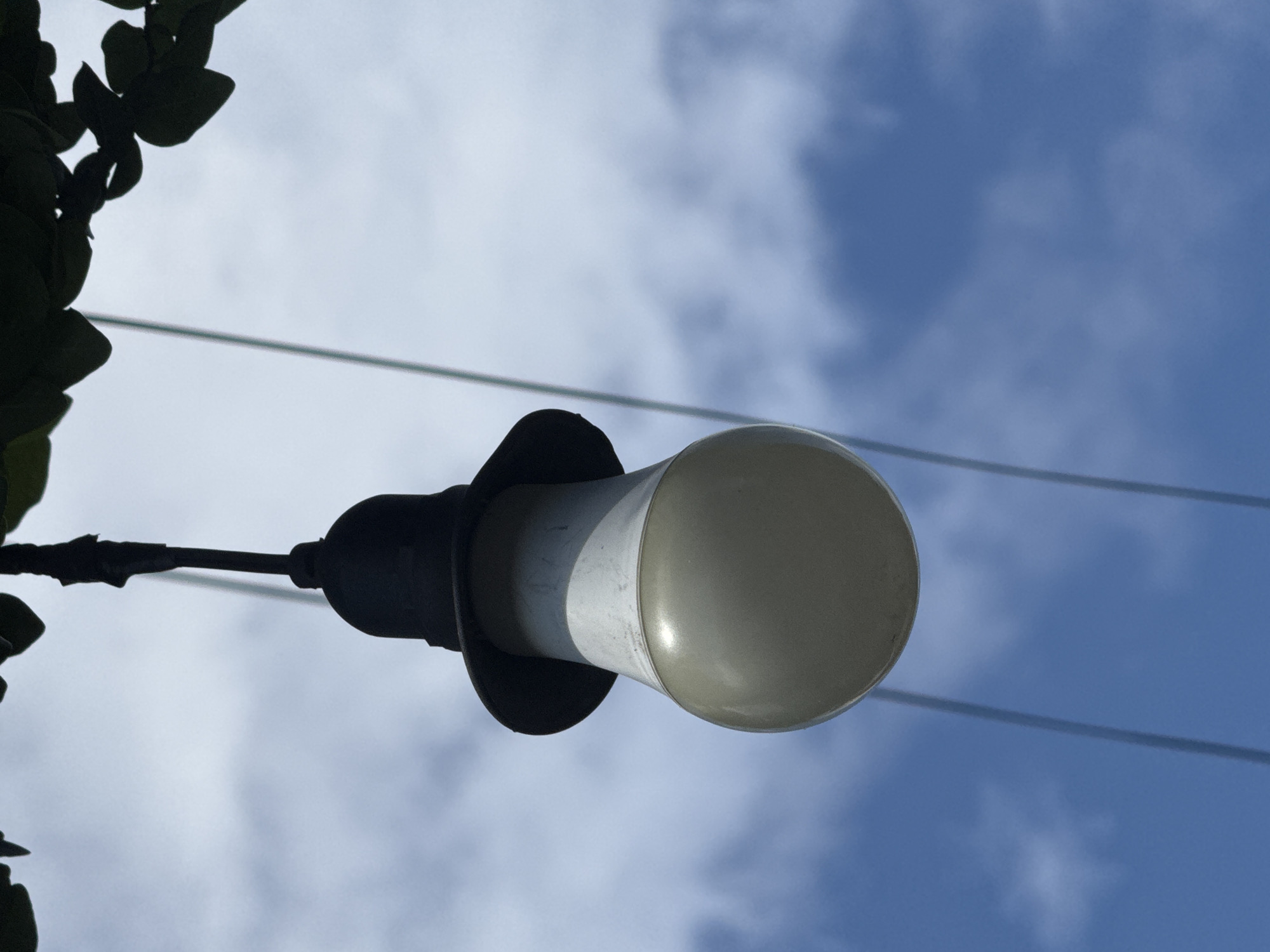 A low-angle shot captures a light fixture against a partly cloudy blue sky. The fixture is attached to a dark-colored, possibly metallic, branch or support structure with green leaves on the left side. The light itself consists of a black base with a flared hood, a white cylindrical section, and a rounded, translucent bulb. Two thin, horizontal wires are visible stretching across the sky in the background. The scene is devoid of people. The location is presumed to be Kuta, Indonesia.